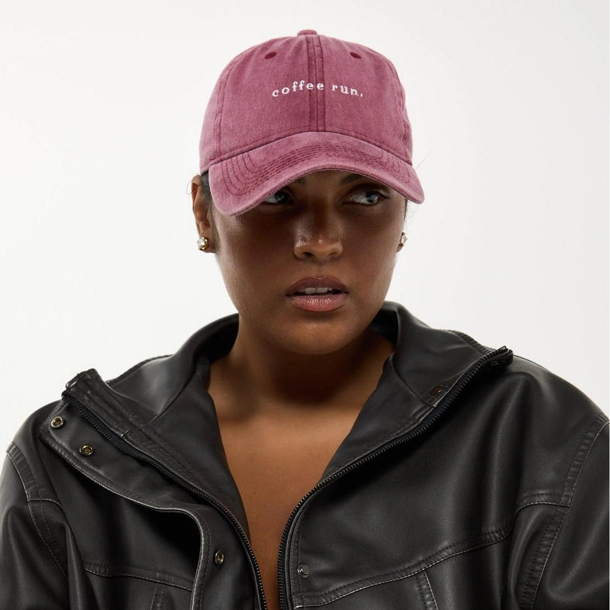 A model wearing a black faux-leather zip-up jacket and a dusty pink baseball cap with 'coffee run.' embroidered on the front, photographed against a plain light background.