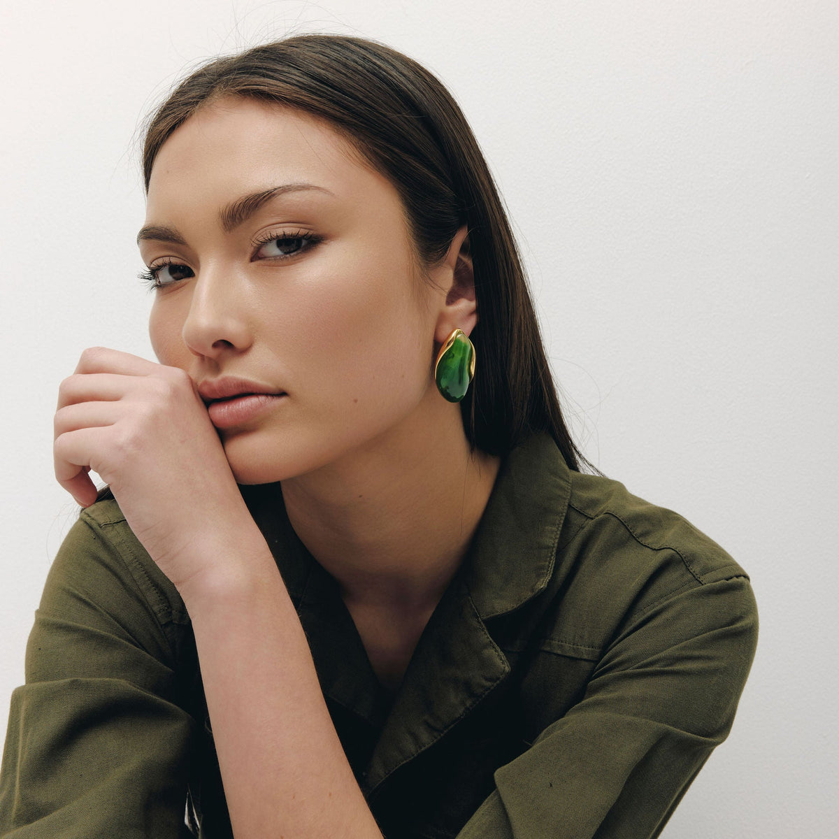 A woman with dark hair wearing a green collared shirt and matching green pants, looking thoughtfully to the side while resting her chin on her hand. She is wearing a prominent green teardrop-shaped earring.