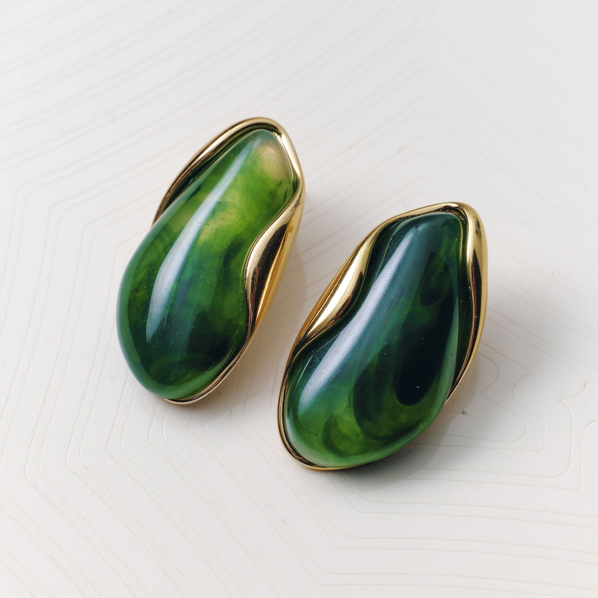 A pair of gold-tone stud earrings with organic, bean-shaped green stone inlays, displayed on a light textured surface.