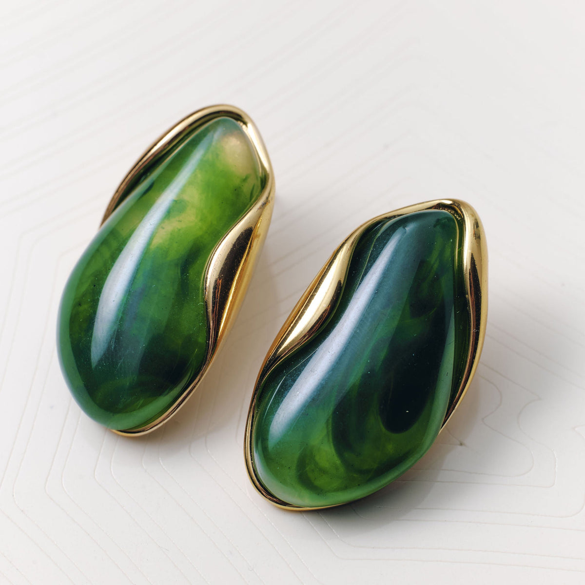 A pair of gold-tone stud earrings with organic, wavy teardrop-shaped green stone inlays, displayed on a light off-white textured surface.