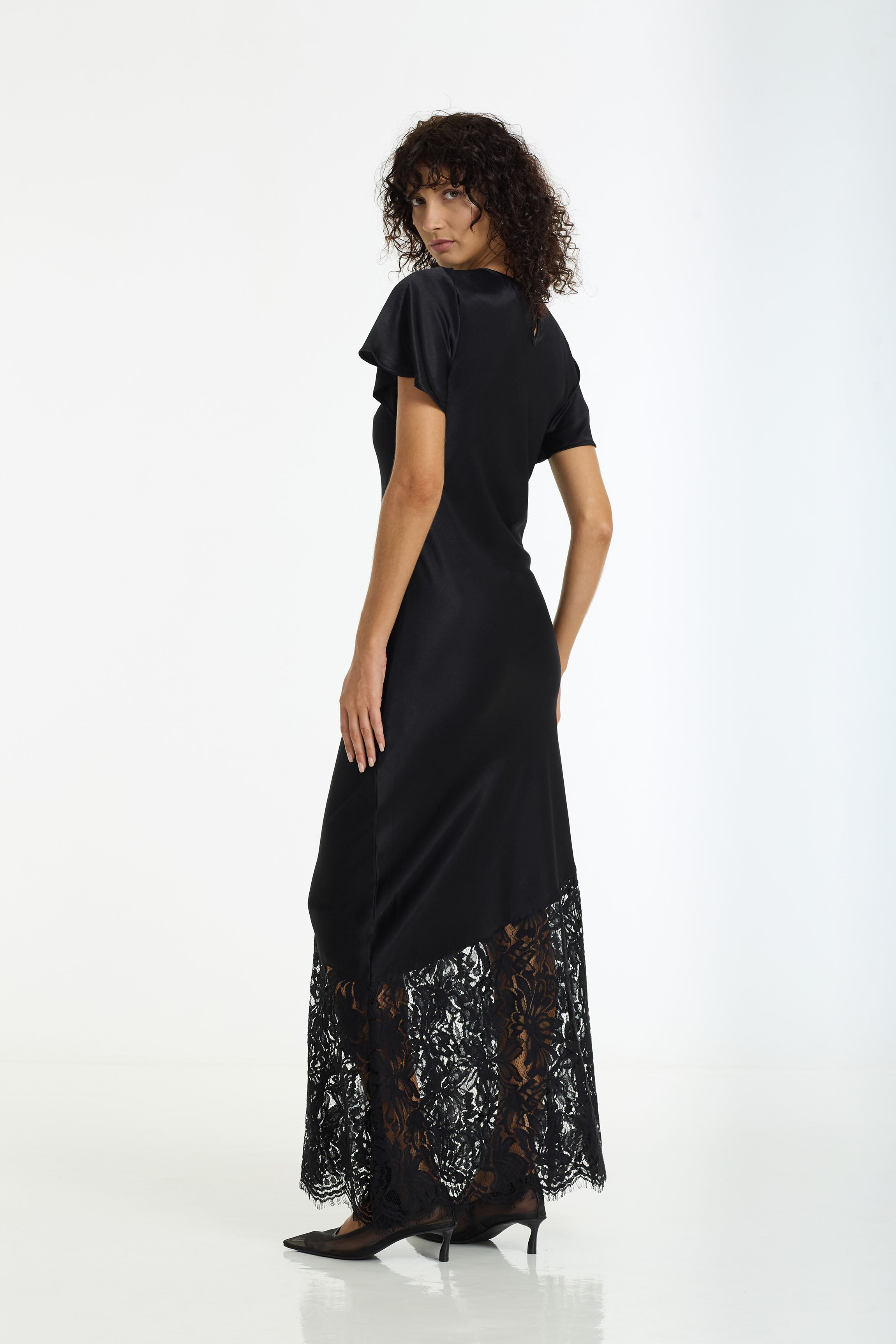 A woman modeling a black maxi dress with short flutter sleeves and a sheer black lace hem, photographed from the back against a plain white studio background.