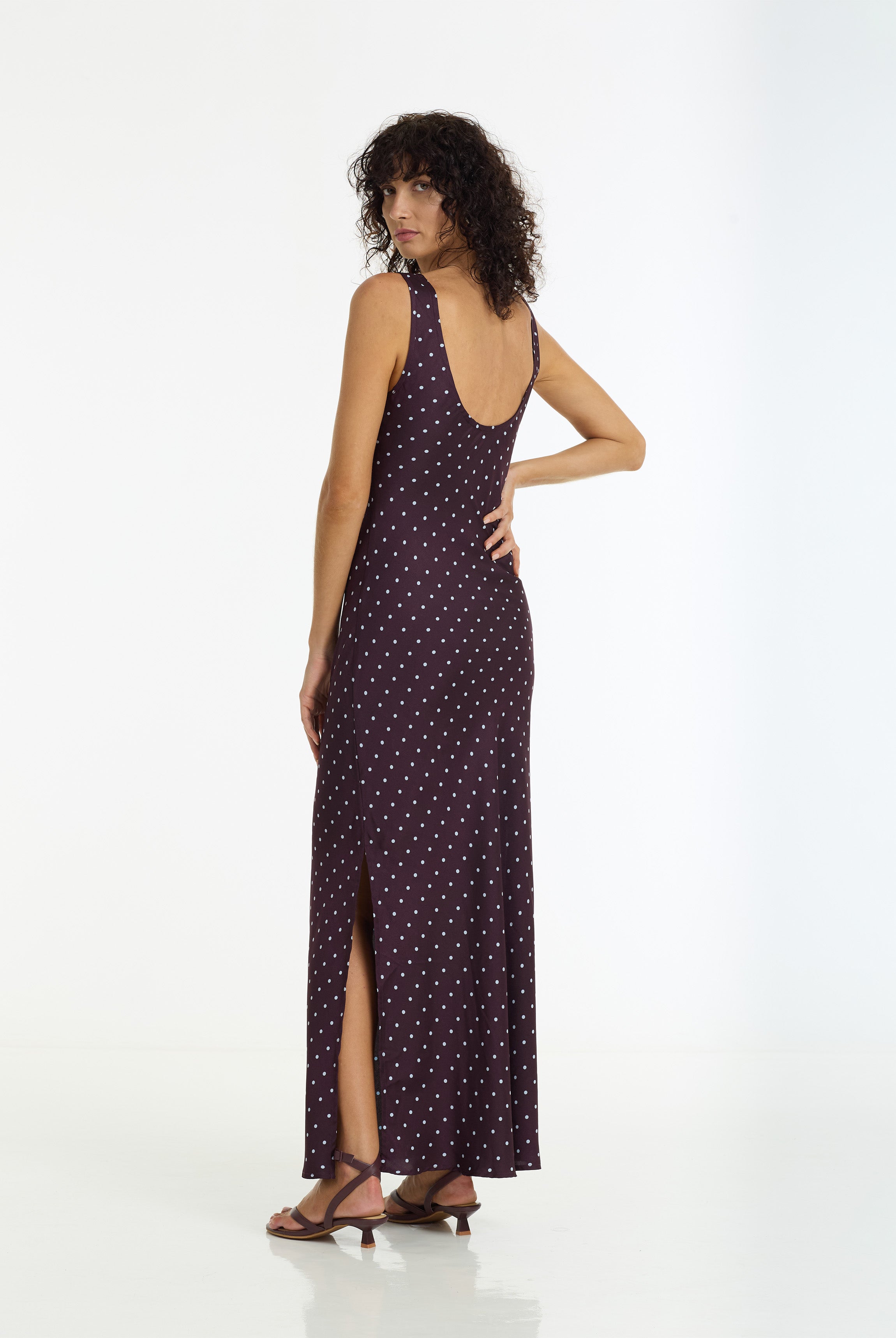 A woman modeling a sleeveless, backless maxi dress with a small white polka dot pattern on a dark purple base, shown from a three-quarters back view against a white studio background.