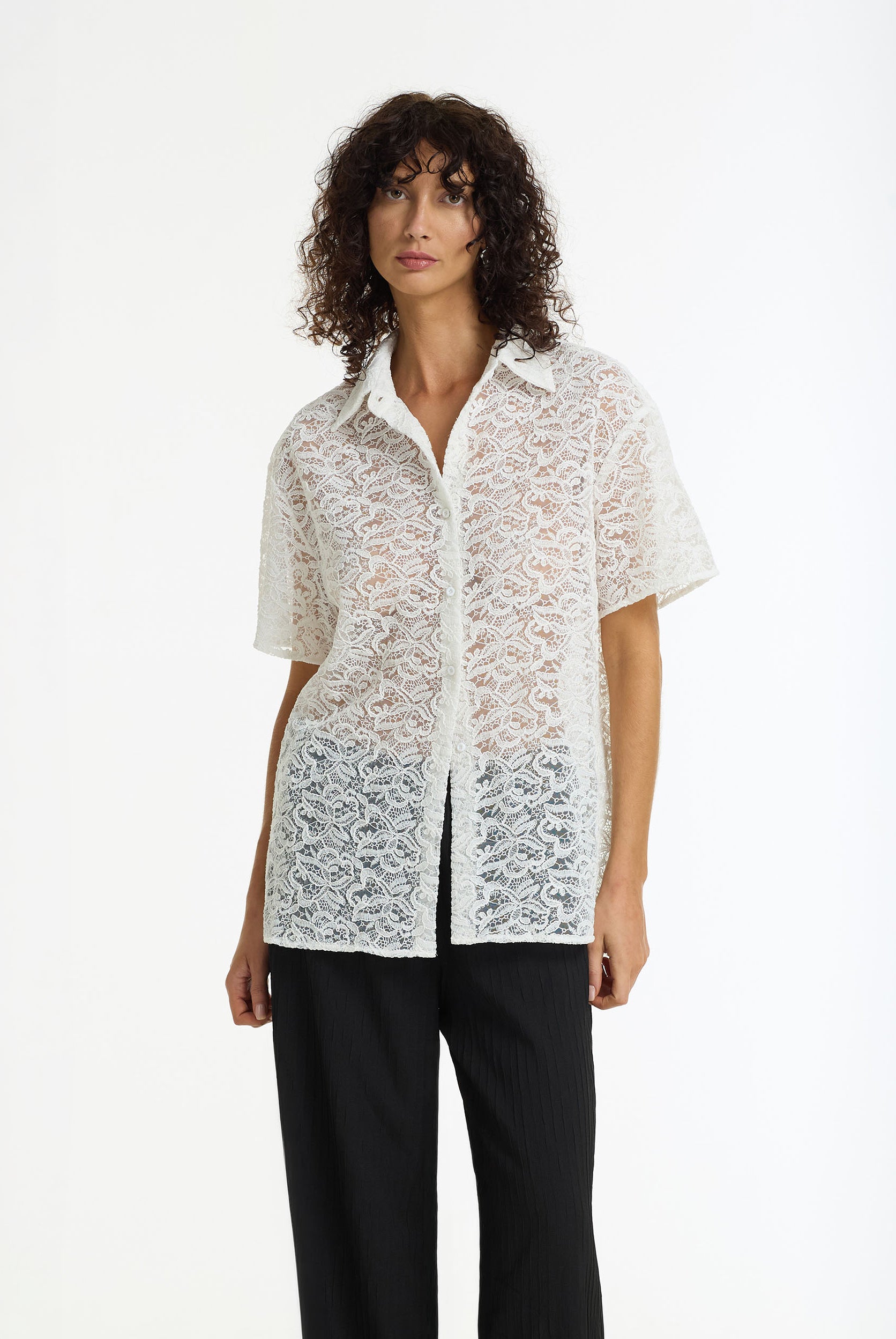 A model with curly dark hair wearing a white short-sleeve lace button-up shirt and black trousers, standing against a plain white studio background.