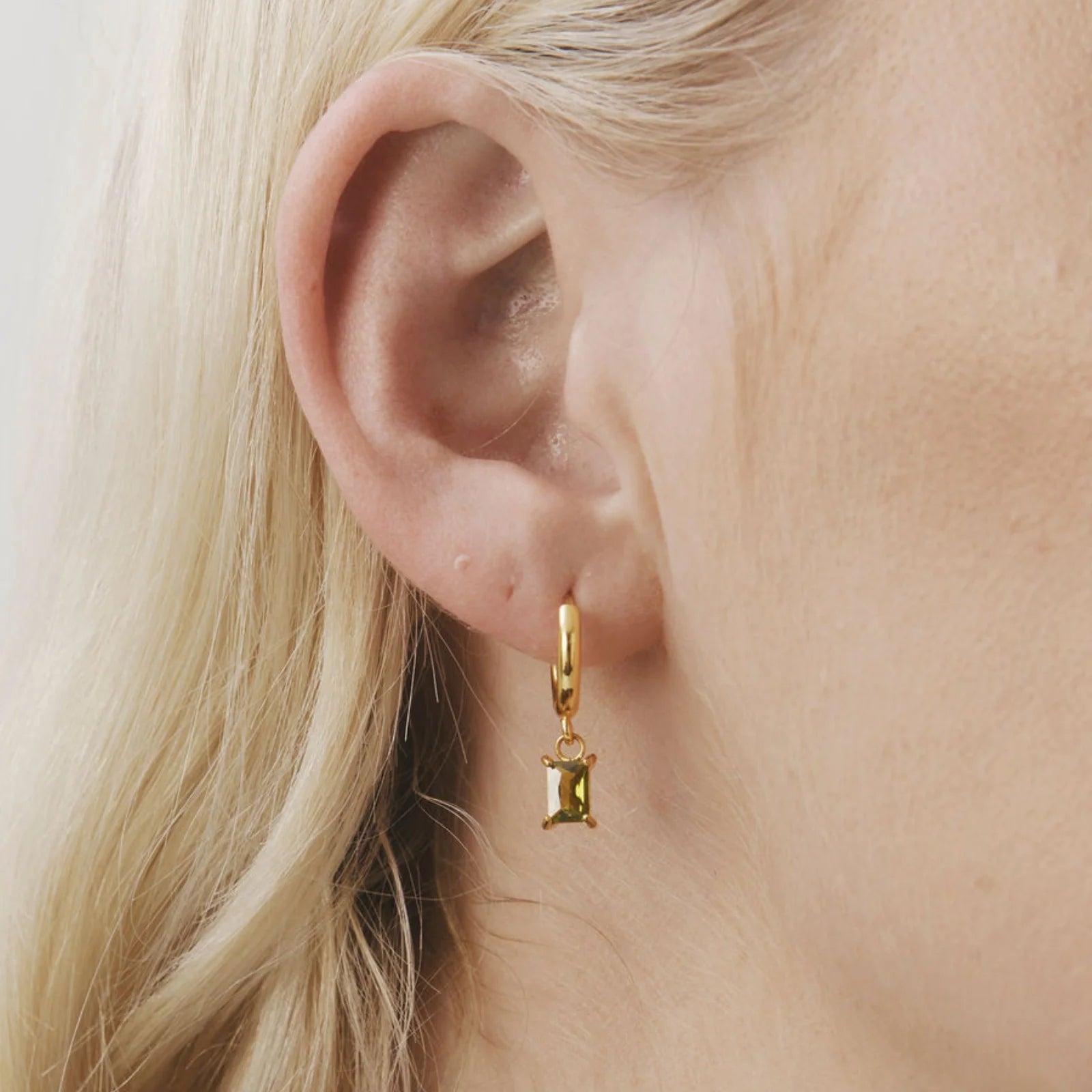 A close-up of a model's ear wearing a delicate gold huggie hoop earring with a small gold hourglass charm.
