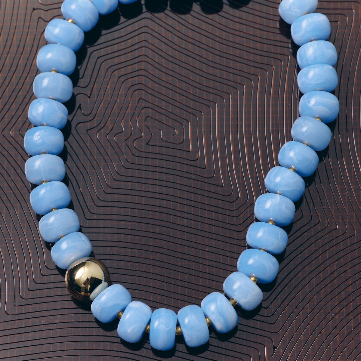 A beaded necklace made of light blue oval beads with a single polished gold accent bead, displayed on a dark brown textured background.