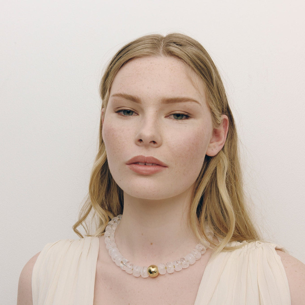 A blonde woman wearing a cream pleated top and a chunky white beaded necklace with a small gold bead accent, photographed against a plain off-white background.