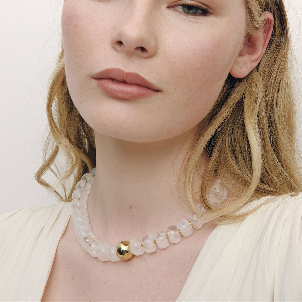 A close-up of a woman wearing a delicate necklace made of translucent pale milky-white beads with a small gold bead accent at the centre, set against a plain light background.