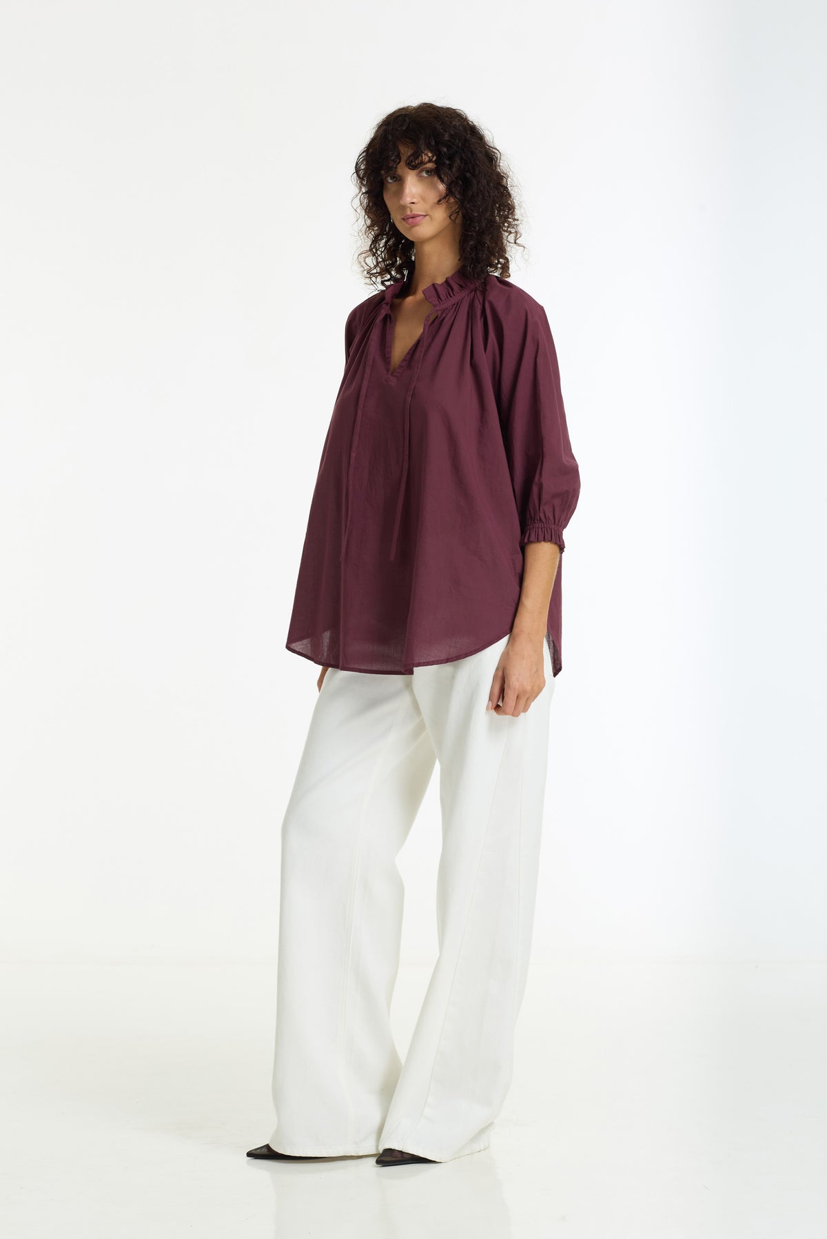 A woman modeling a loose-fitting, deep burgundy blouse with three-quarter sleeves and a split neckline, paired with wide-leg white trousers, against a plain white studio background.