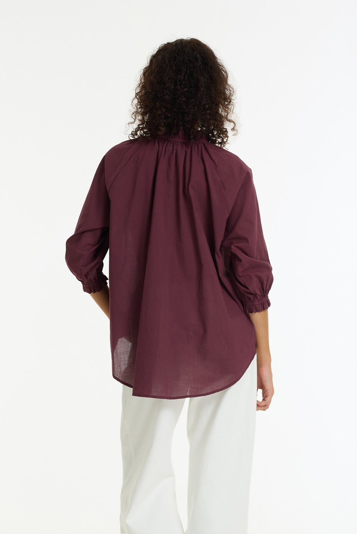 A woman viewed from the back wearing a loose, gathered long-sleeve blouse in deep burgundy with white pants, set against a plain white studio background.