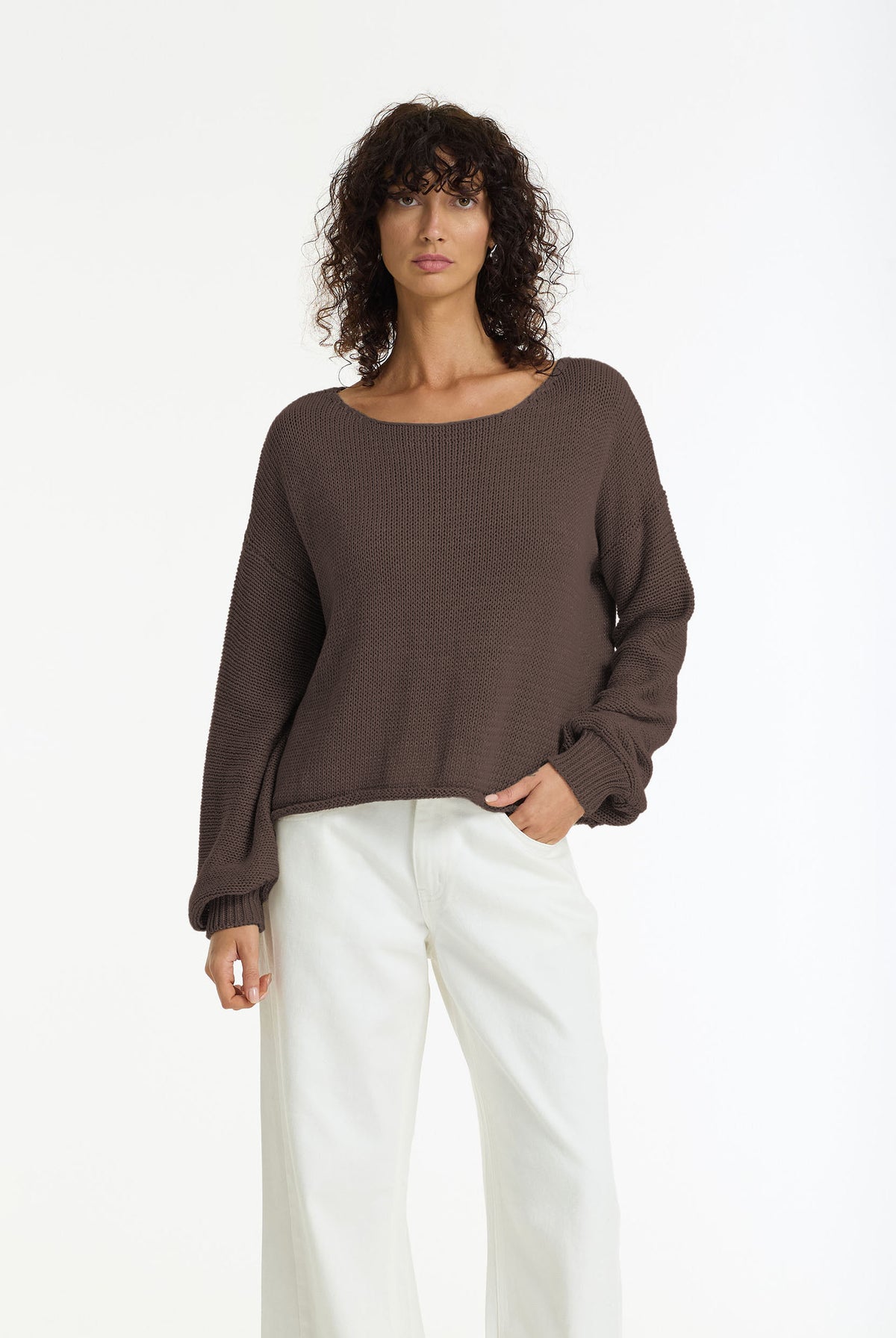 A woman modeling a Close Cotton Yarn Sweater in Chocolate by BLAK, paired with white wide-leg pants, against a plain white studio background.