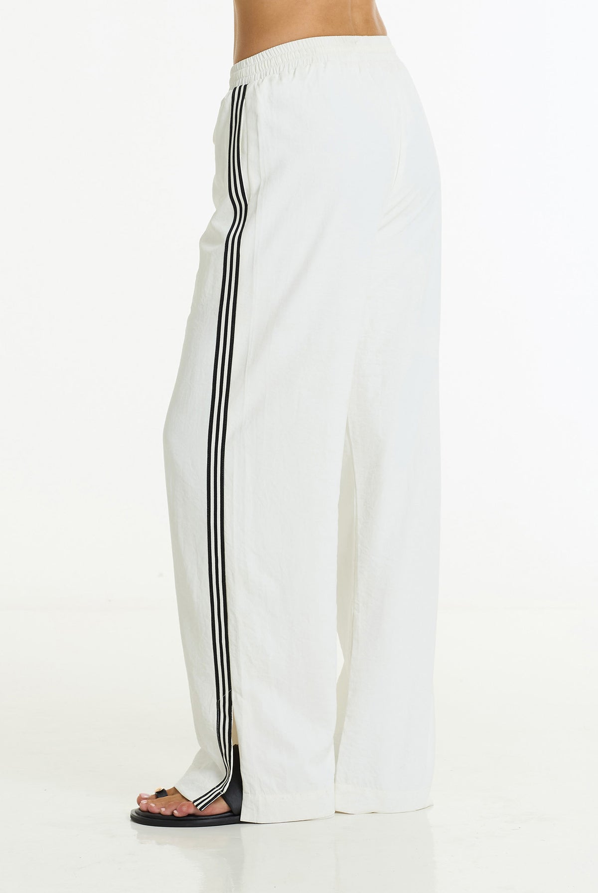 White wide-leg track pants with black double stripes down the side, shown on a model from the waist down against a white studio background.