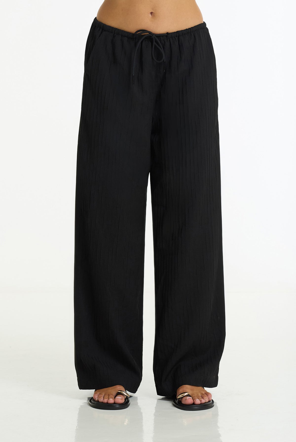 Black wide-leg pants with a drawstring waist, shown on a model from the waist down wearing black sandals against a white studio background.