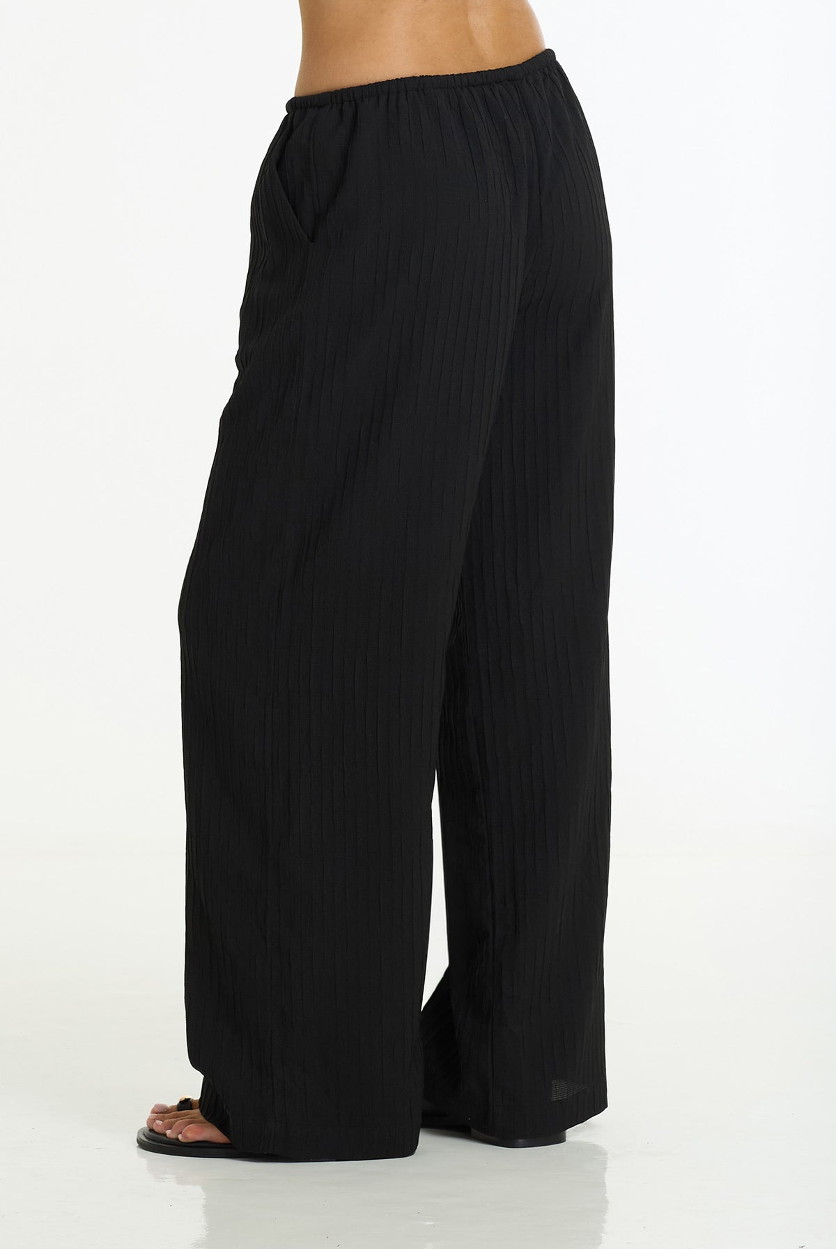 Black wide-leg pleated pants shown on a model from the side, featuring a gathered elastic waistband and side pockets, set against a plain white studio background.