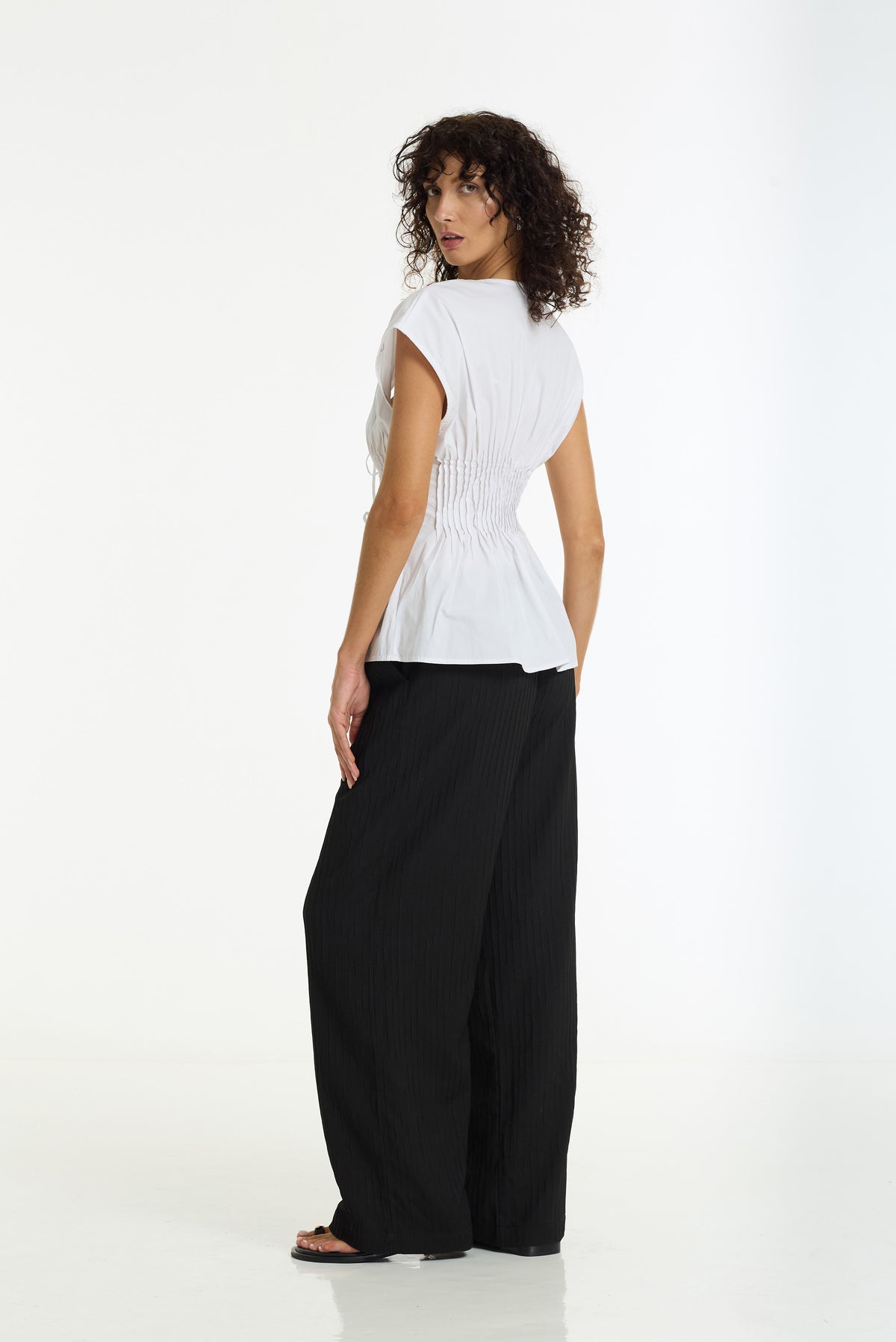 A woman with curly dark hair is shown from the back wearing a white sleeveless top with a gathered peplum-like back and black wide-leg pleated pants, standing against a plain white studio background.