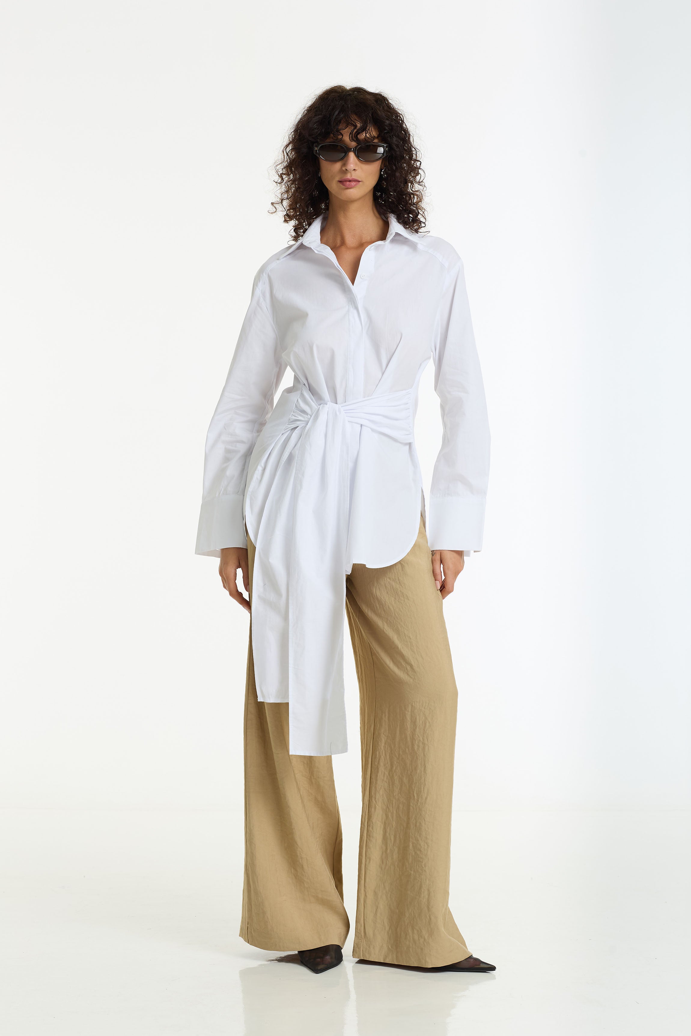 A model wearing a white long-sleeve button-up shirt tied at the waist with the Eternity Wide Leg Tailored Pant in camel by BLAK, styled with dark sunglasses and black pointed shoes against a plain white studio background.