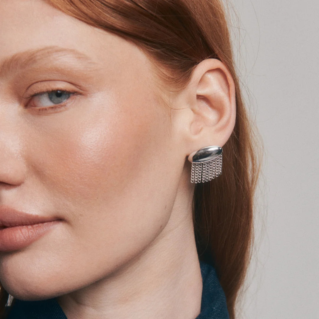A close-up of a woman's ear wearing a silver geometric stud earring with a textured, woven-like lower section and a smooth dome on top, set against a neutral gray background.