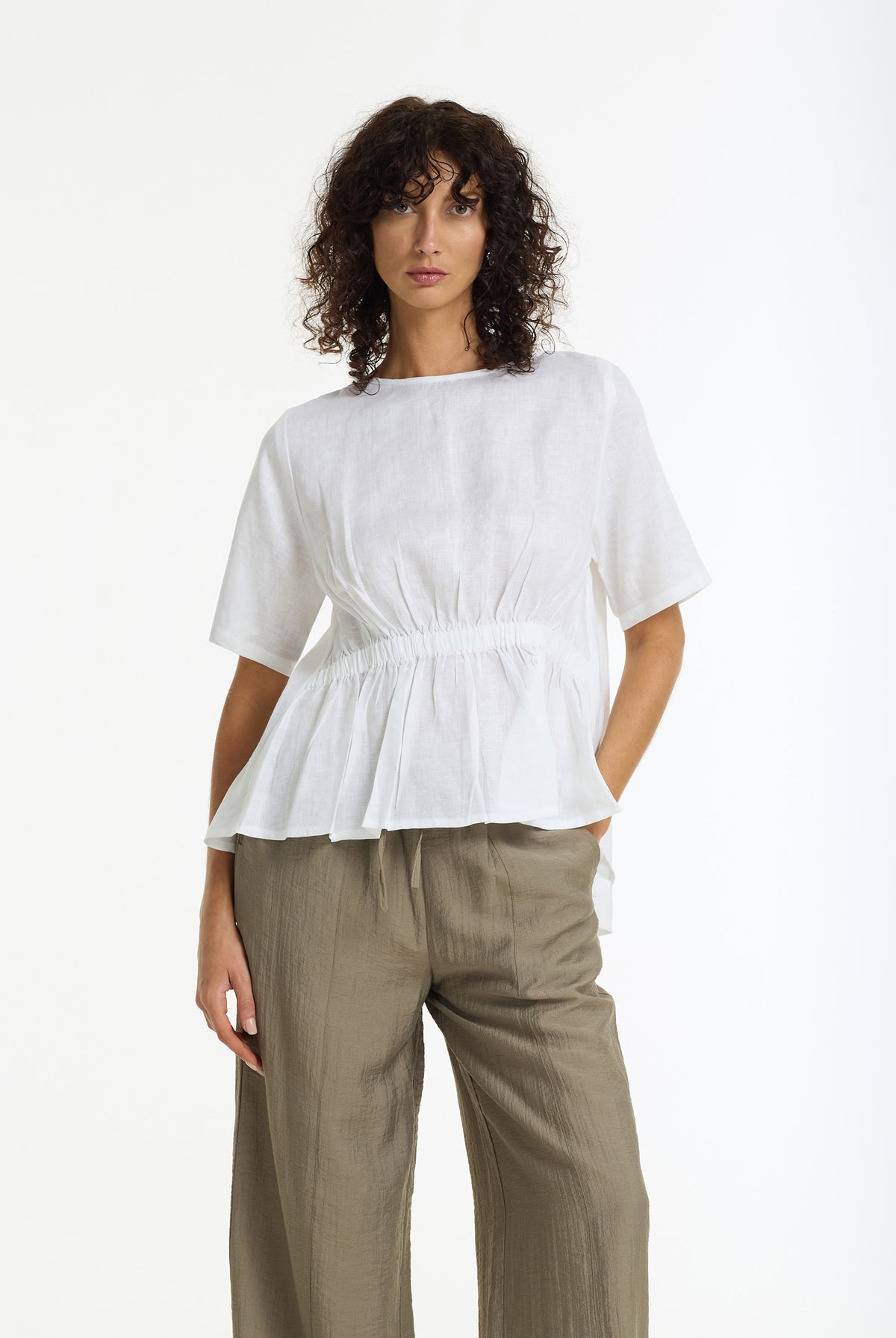 A model wearing a Gentle Blossom Linen Top in White by BLAK and olive green wide-leg trousers, standing against a plain white studio background.