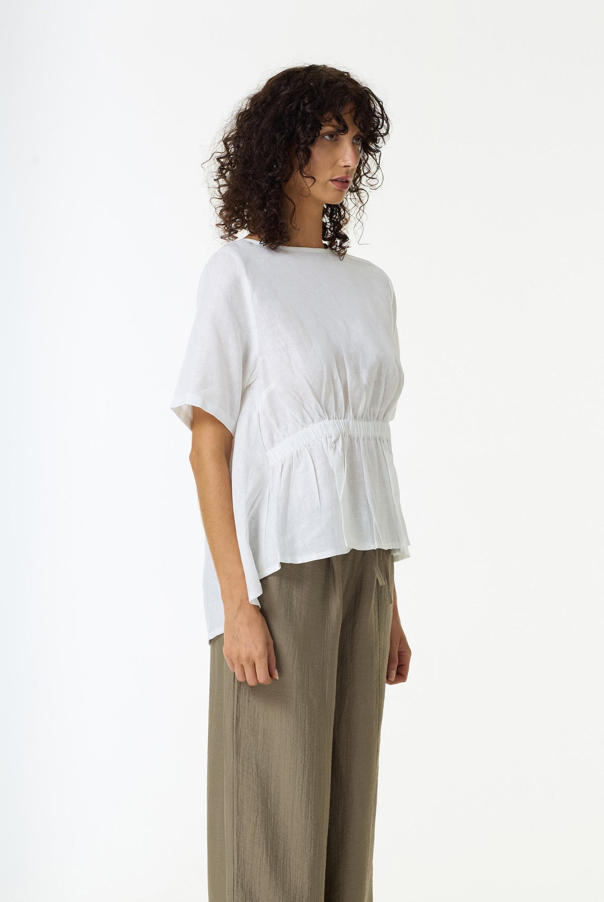 A woman with curly dark hair models a white short-sleeve linen top with a gathered waist and olive green wide-leg trousers against a plain white background.