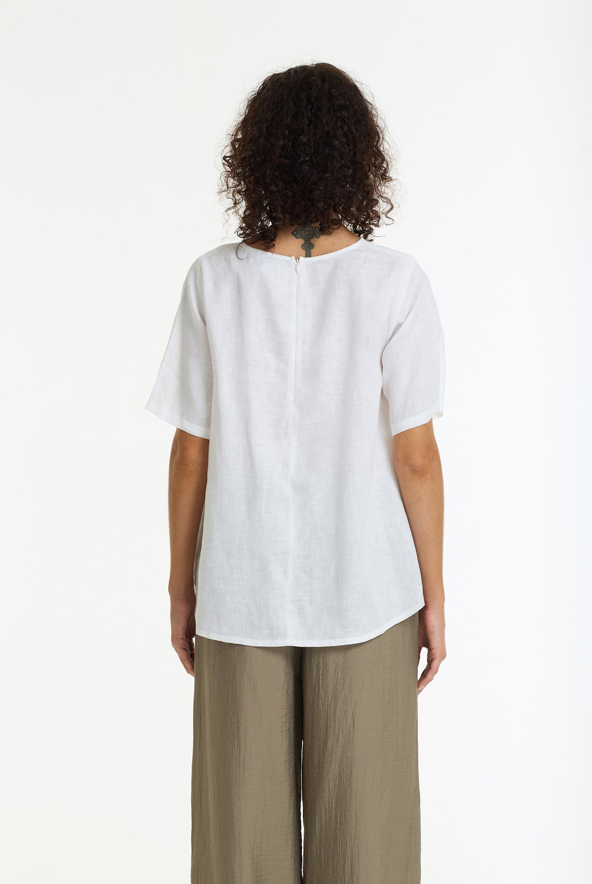 A woman viewed from the back wearing a loose white short-sleeve top and olive green wide-leg pants, photographed against a plain white studio background.