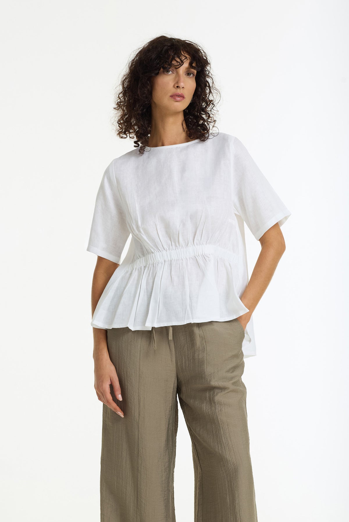 A woman modeling a white short-sleeve top with a gathered waist and peplum hem, paired with olive green wide-leg trousers, against a plain white studio background.