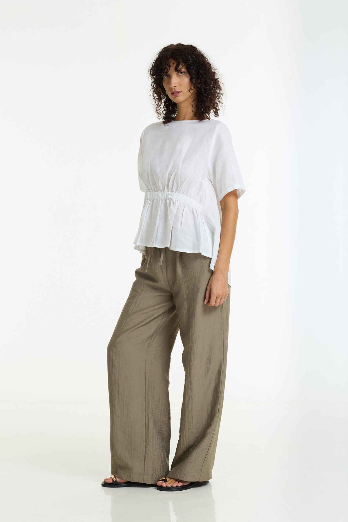 A woman modeling a white short-sleeve linen peplum top and wide-leg taupe pants against a clean white studio background.
