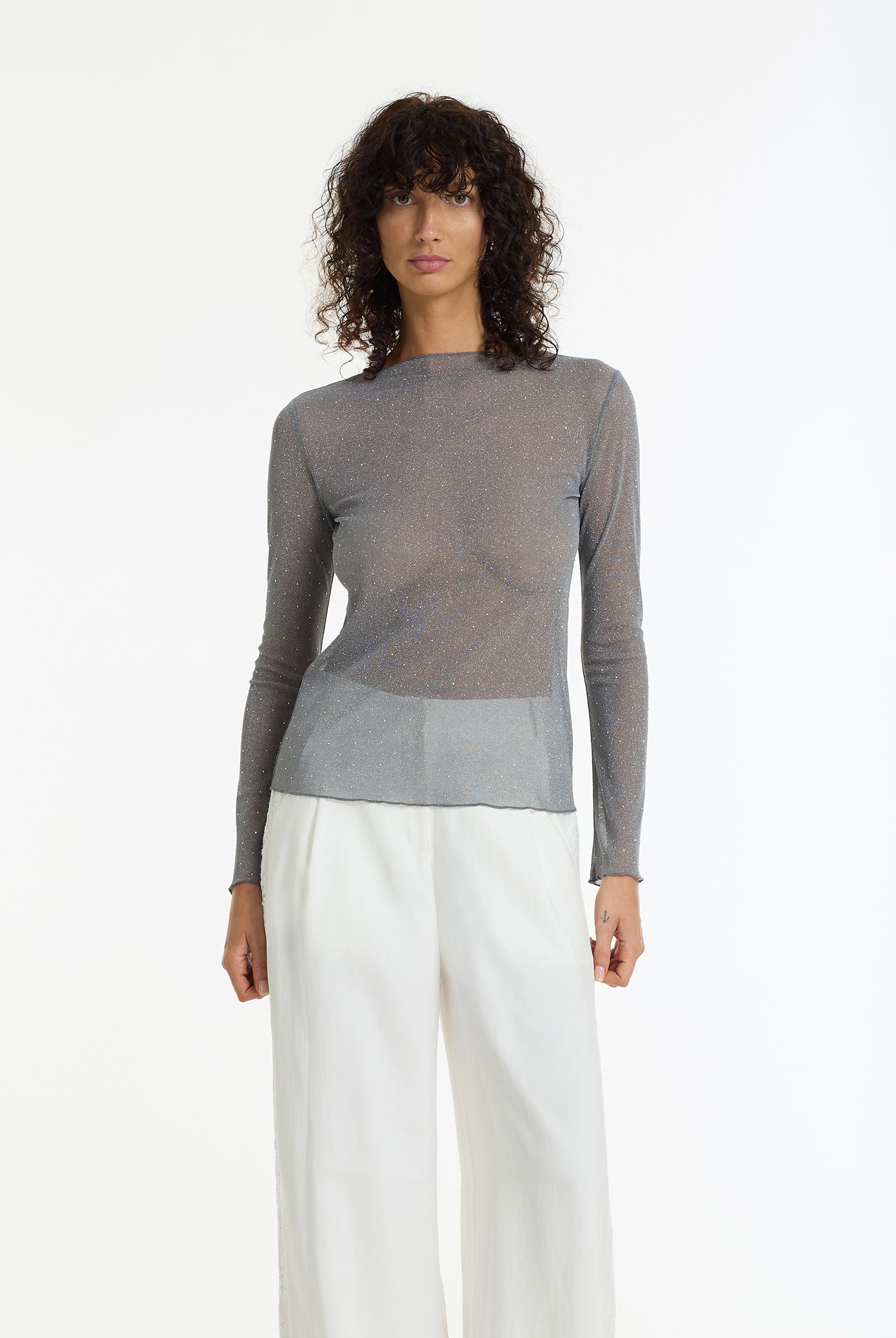 A person wearing the Gigi Diamanté Long Sleeve Top by BLAK, a charcoal sheer stretch top with silver diamantés, long sleeves and a softly fitted silhouette and white pants on a white background.