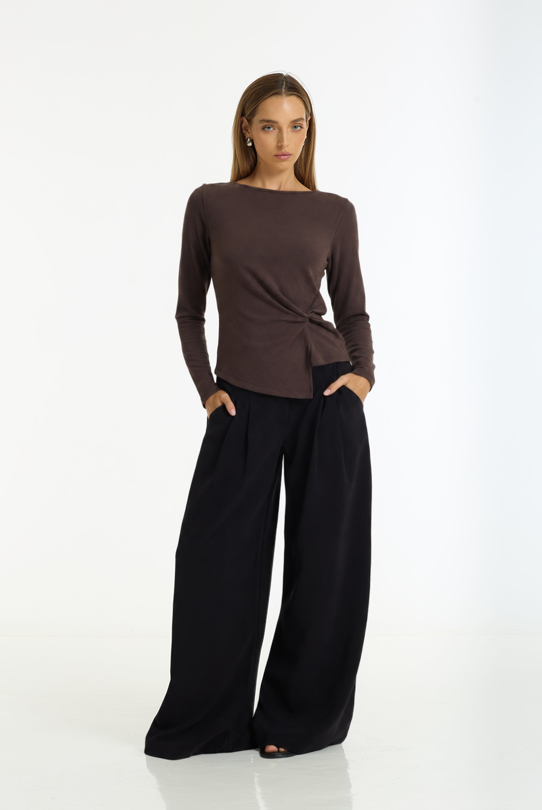 A woman modeling a dark brown long-sleeve top with a twisted front detail and black wide-leg pants, standing against a plain white studio background.