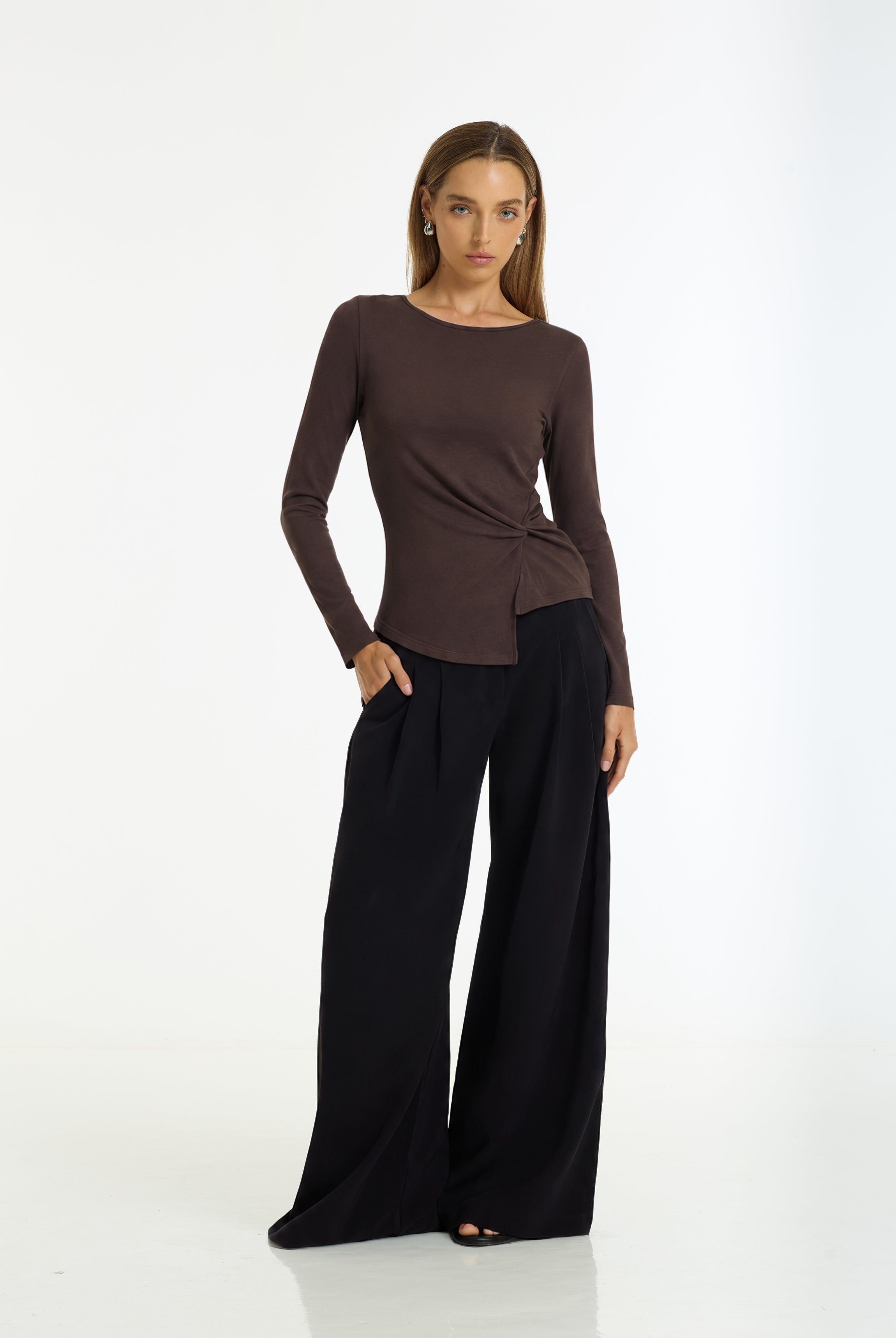 A woman modeling a dark brown long-sleeve top with a twisted side detail and black wide-leg pants against a plain white studio background.