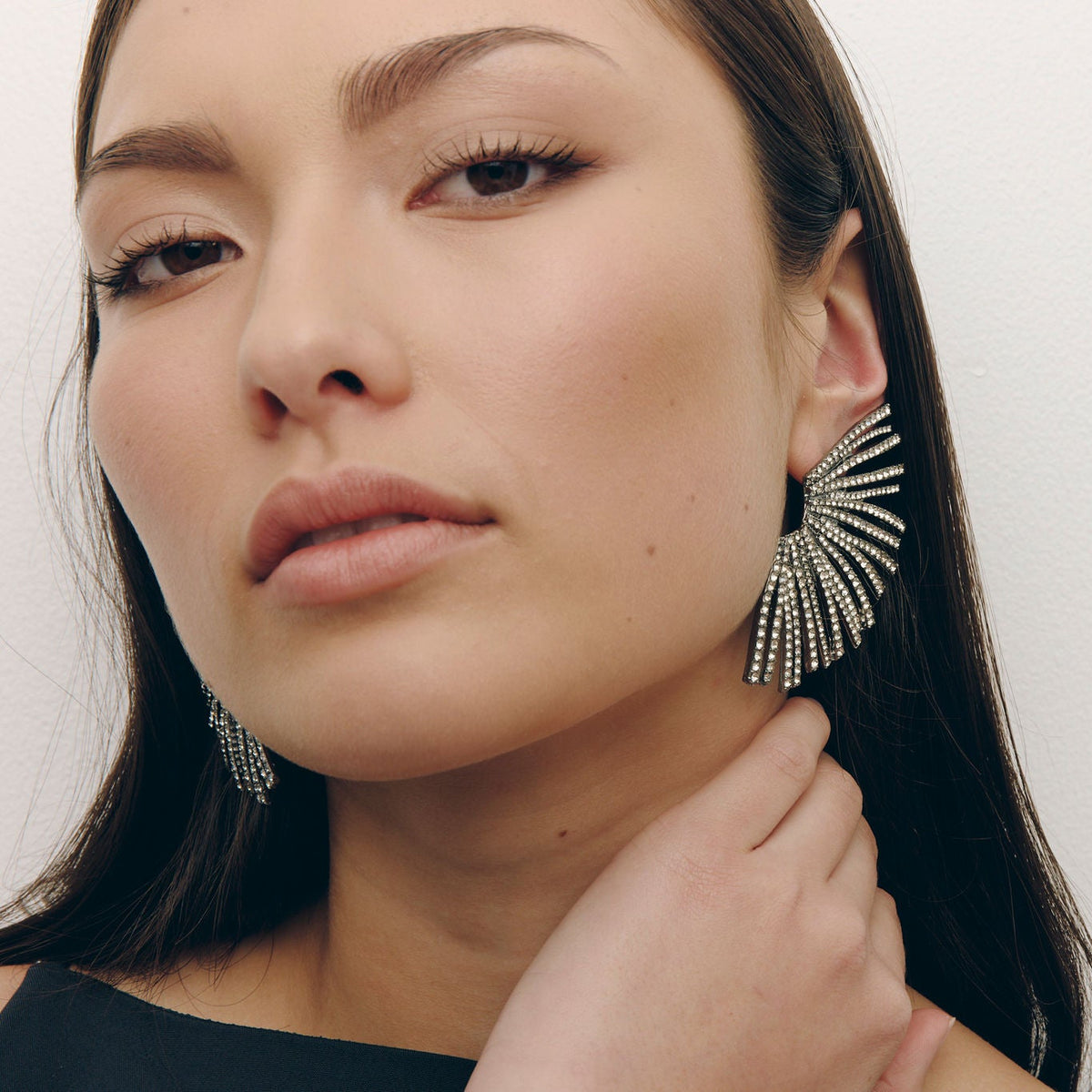 A close-up of a woman wearing large silver statement earrings with a sunburst or fan-like design made of small dangling stones, against a plain light background.