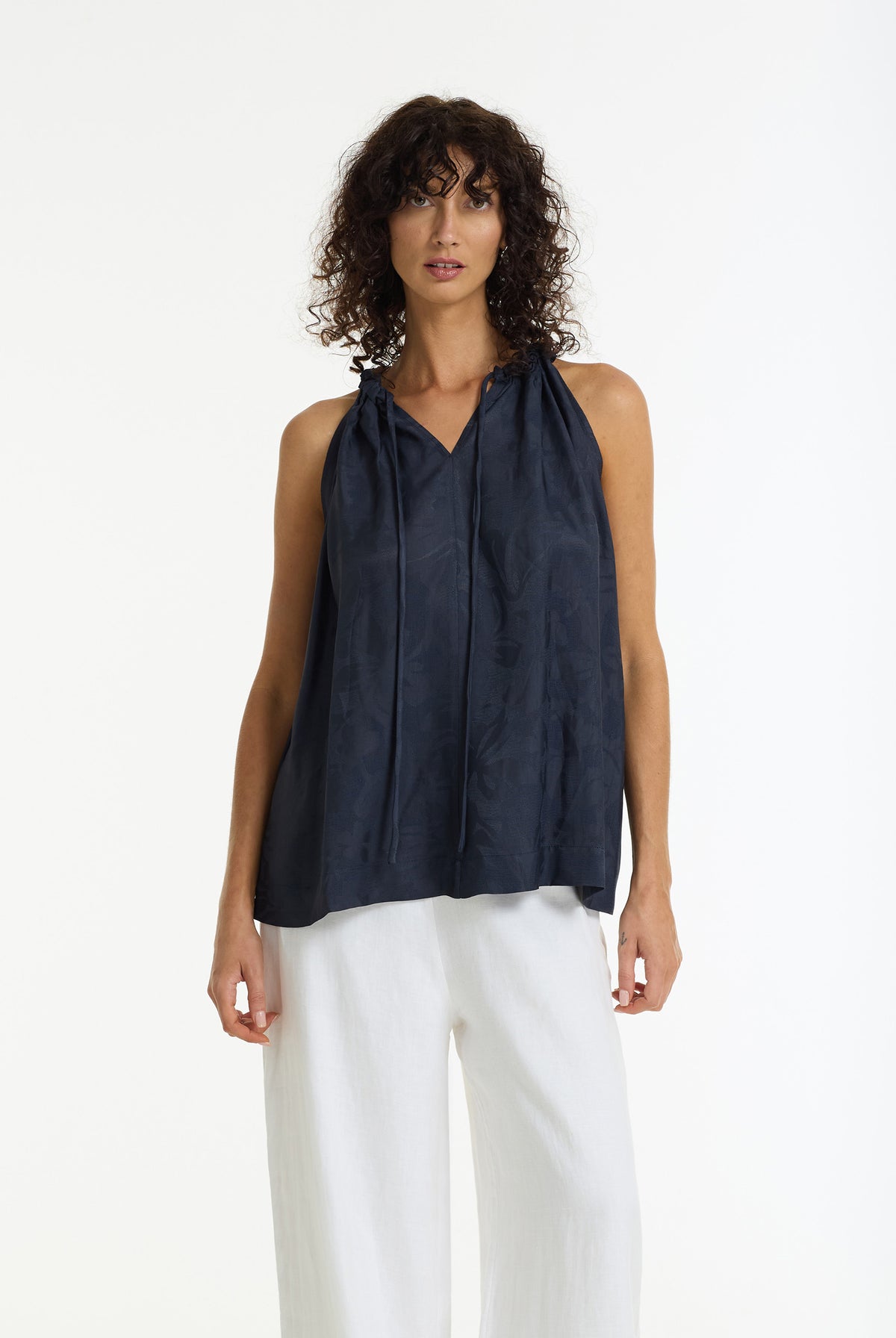 A model with curly dark hair wearing a Meet Me Halter Top in Dark Indigo by BLAK and white wide-leg trousers, standing against a plain white studio background.