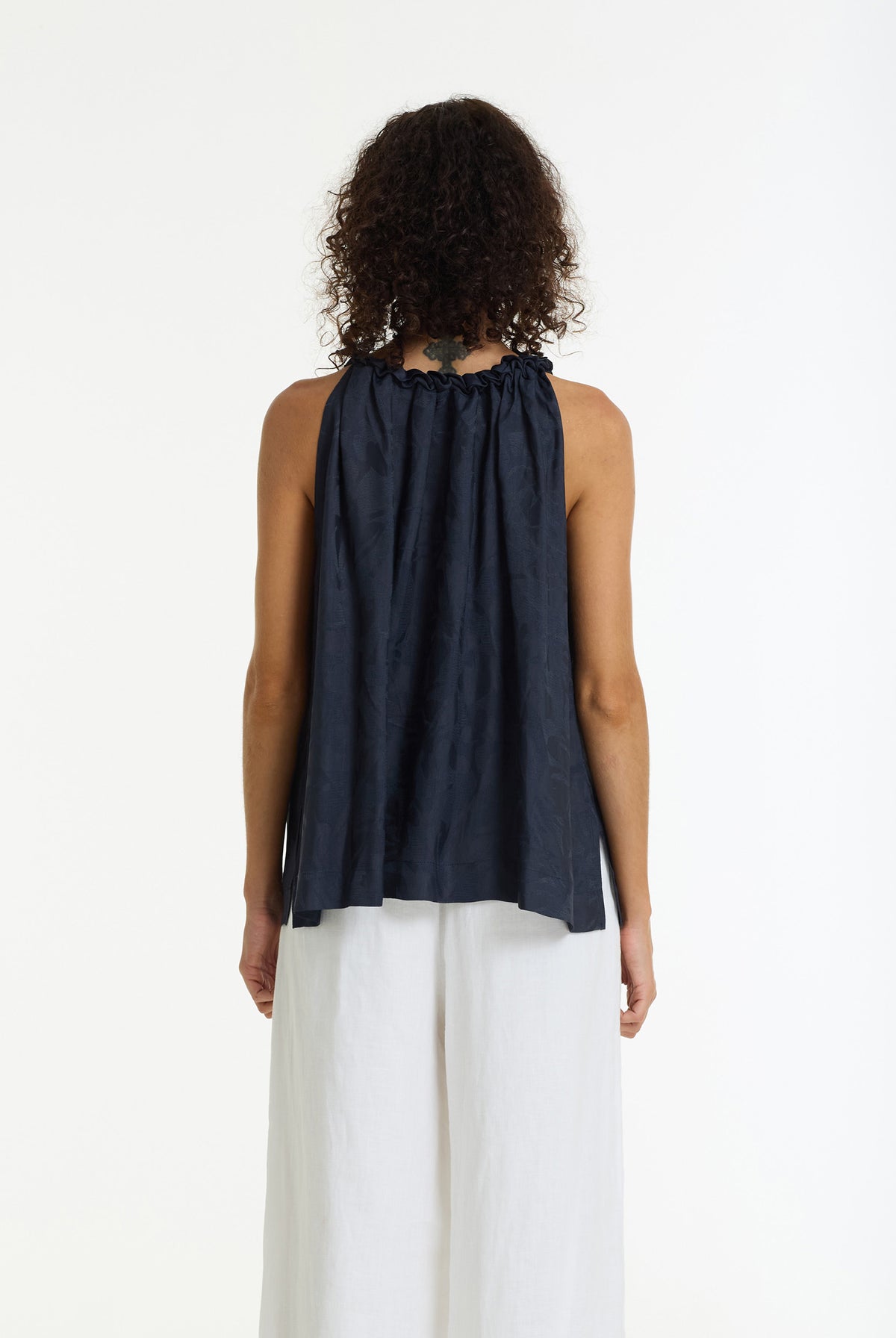 A woman viewed from the back wearing a sleeveless dark navy top with a gathered neckline and white wide-leg pants, against a plain white studio background.