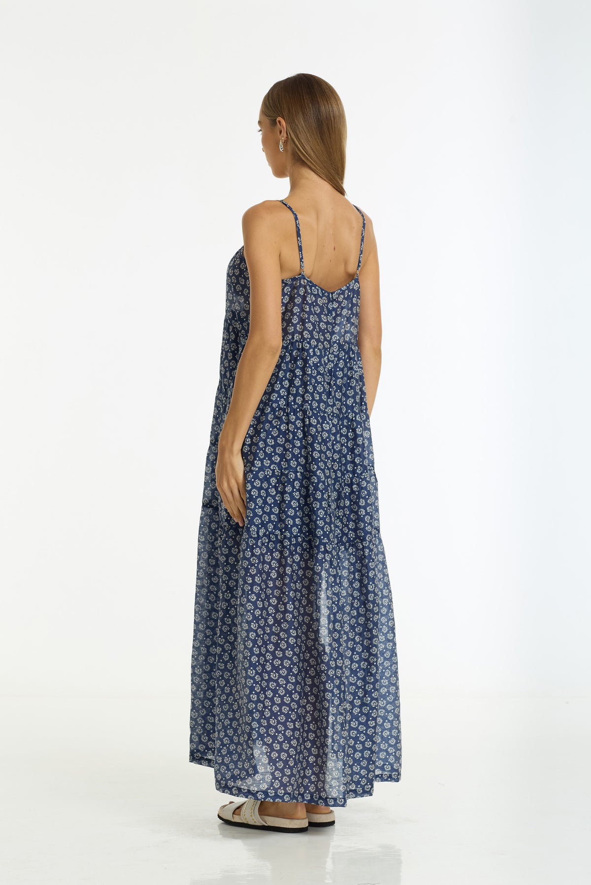 A woman viewed from the back wearing a sleeveless, floor-length blue maxi dress with a small white floral pattern and thin shoulder straps, standing against a plain white studio background.