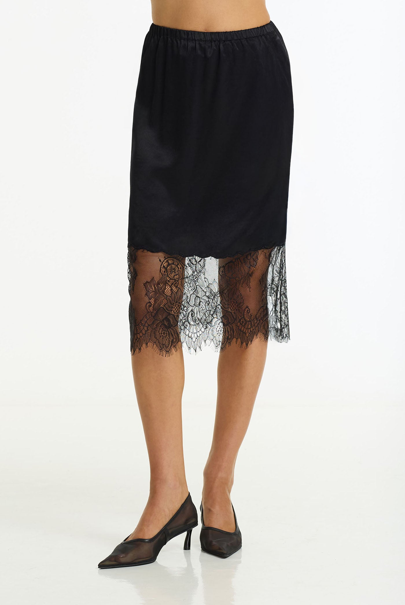 A black satin slip skirt with a sheer black lace hem, modeled on a person wearing black pointed-toe heels against a plain white studio background.