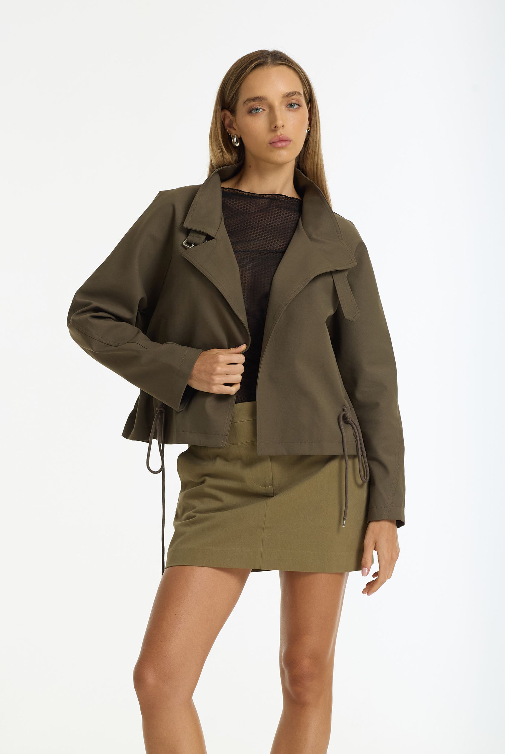 A model wearing a Rhodes Cropped Utility Jacket in dark khaki by BLAK over a Black Gigi Top and a olive green mini skirt, shot against a plain white studio background.