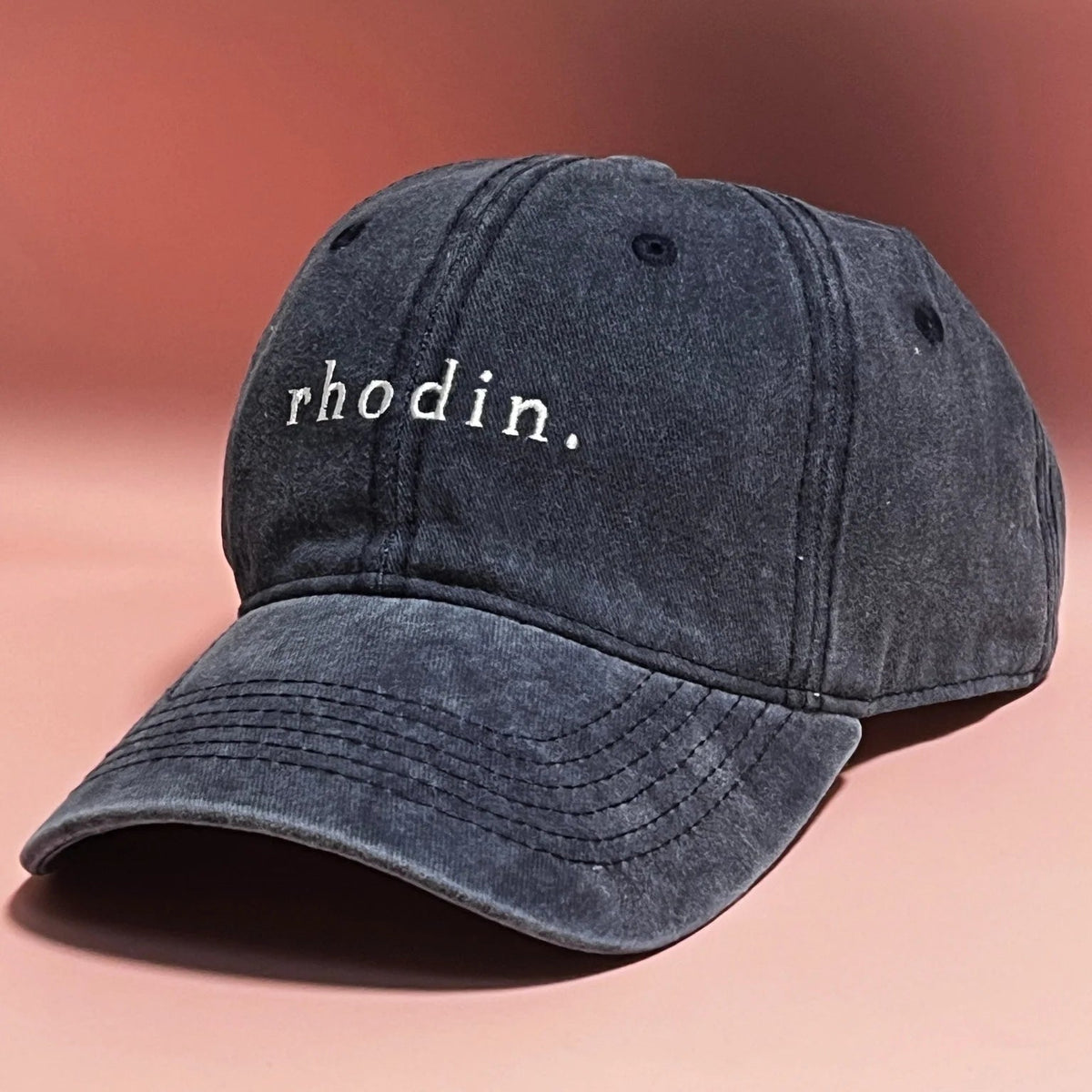 A dark washed denim baseball cap with the word 'rhodin.' embroidered in white on the front, set against a warm pinkish-brown gradient background.