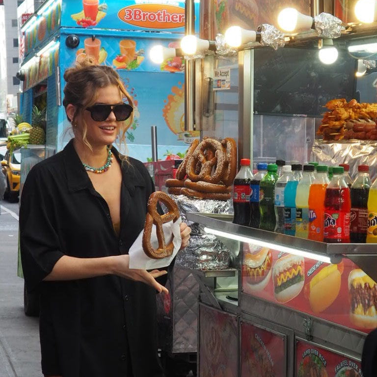 A woman in a black shirt and sunglasses stands at a street food cart holding a pretzel, with hot dogs, drinks, and a blue 3Brother food truck in the background.