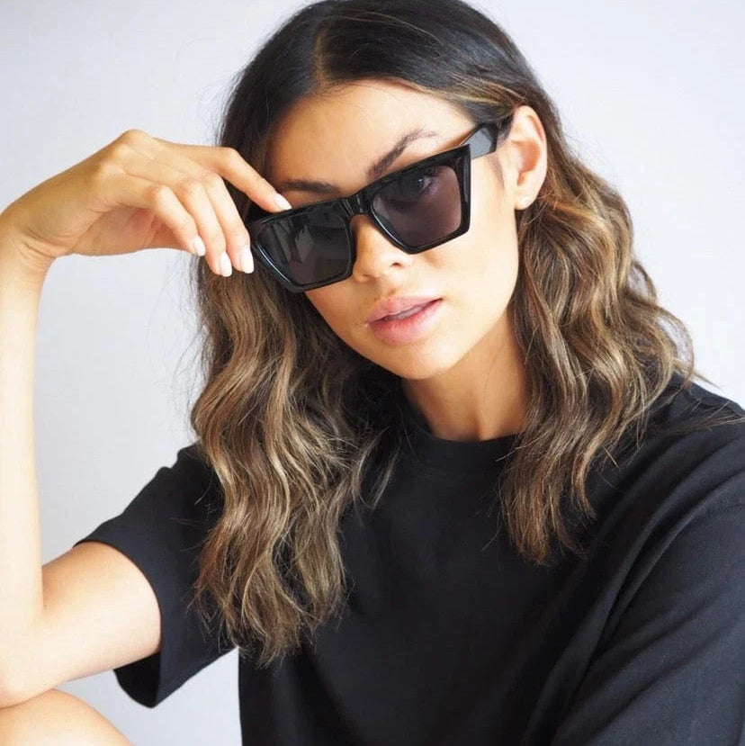 A woman with wavy brown hair wearing black square sunglasses and a black t-shirt, posing against a light gray background.