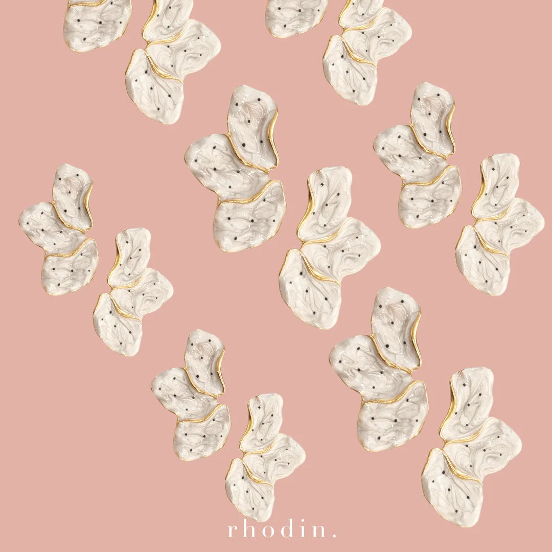 Twelve white rhinestone-embellished butterfly-shaped earrings with gold trim, arranged on a solid dusty pink background with the brand name 'rhodin.' printed at the bottom.