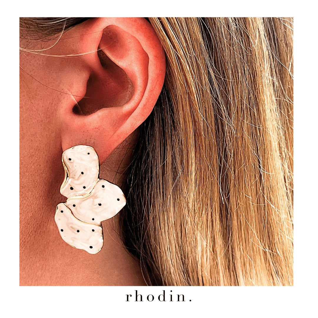 A close-up of a person's ear wearing a cluster of organic-shaped earrings in a warm off-white color with small black polka dots. The image has a white border with the brand name 'rhodin.