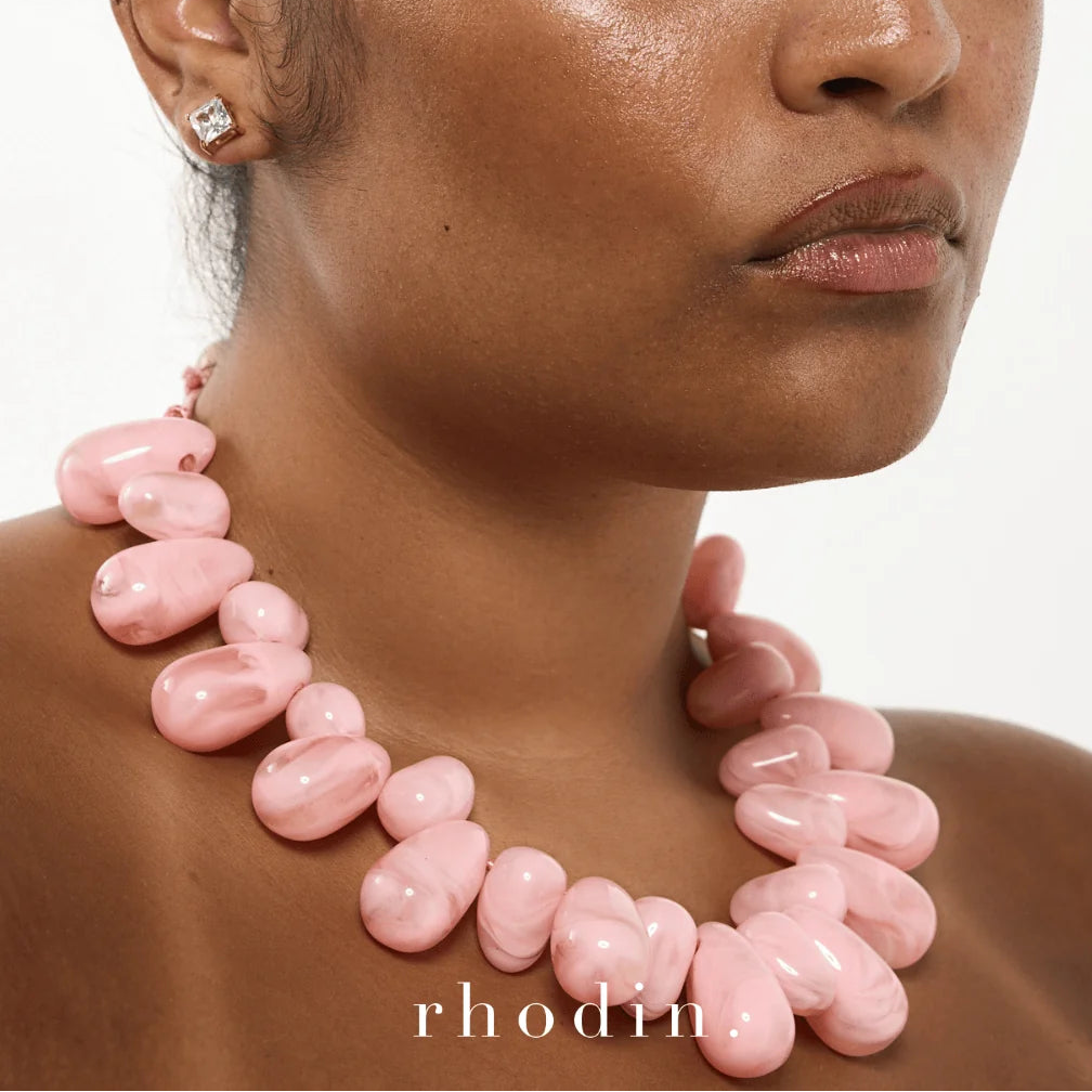 A close-up of a model wearing a chunky, organic-shaped light pink resin necklace by Rhodin, set against a clean white background.