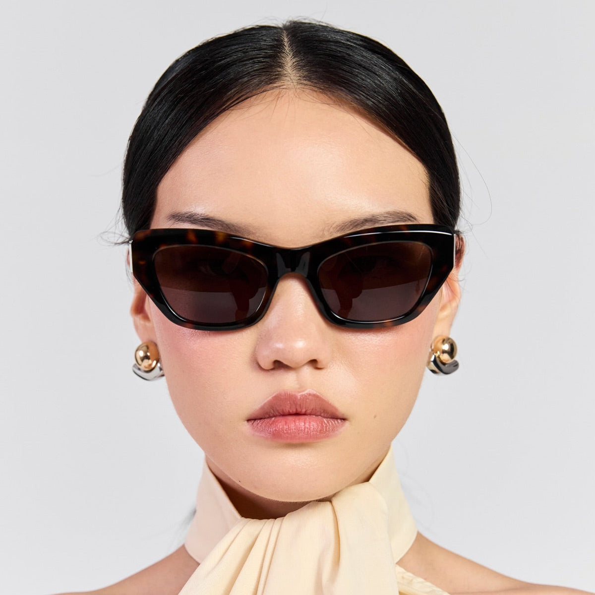 A close-up portrait of a model wearing black cat-eye sunglasses with dark lenses and gold-toned earrings, paired with a cream halter-neck top, against a light gray background.