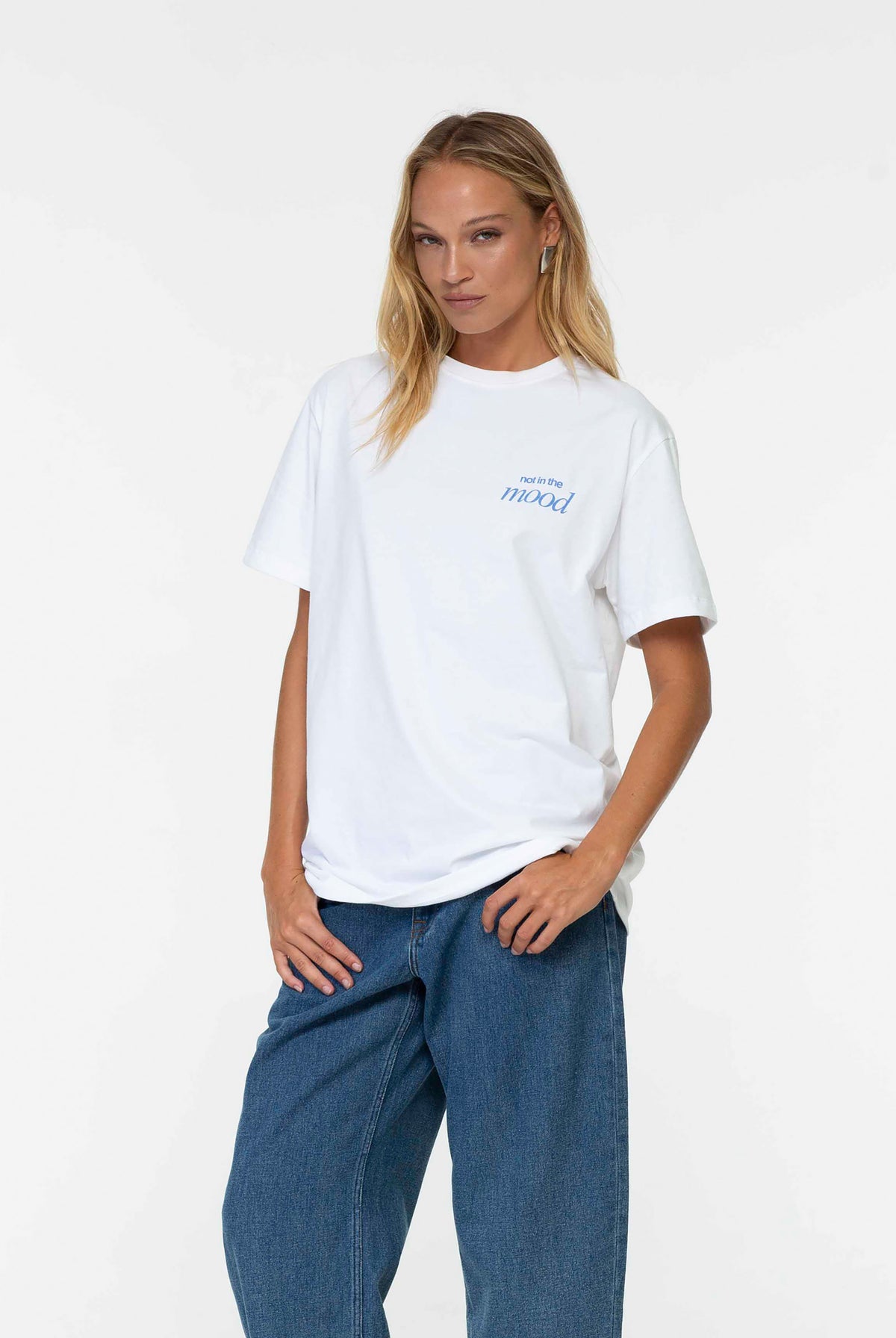 A woman modeling a white short-sleeve t-shirt with a small blue graphic on the left chest, paired with loose blue jeans, against a plain white studio background.