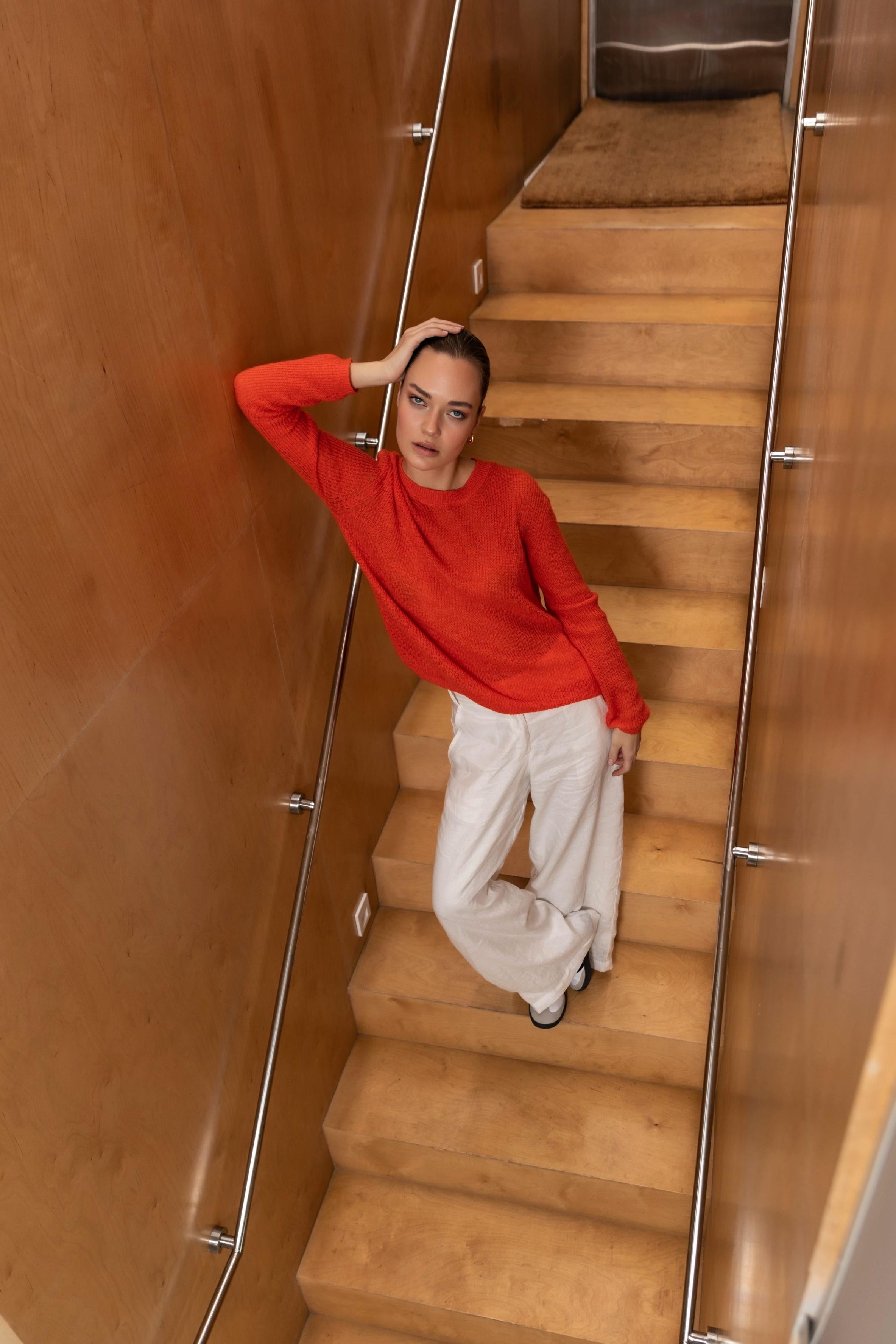 Person in a orange sweater and white pants standing on a wooden staircase.