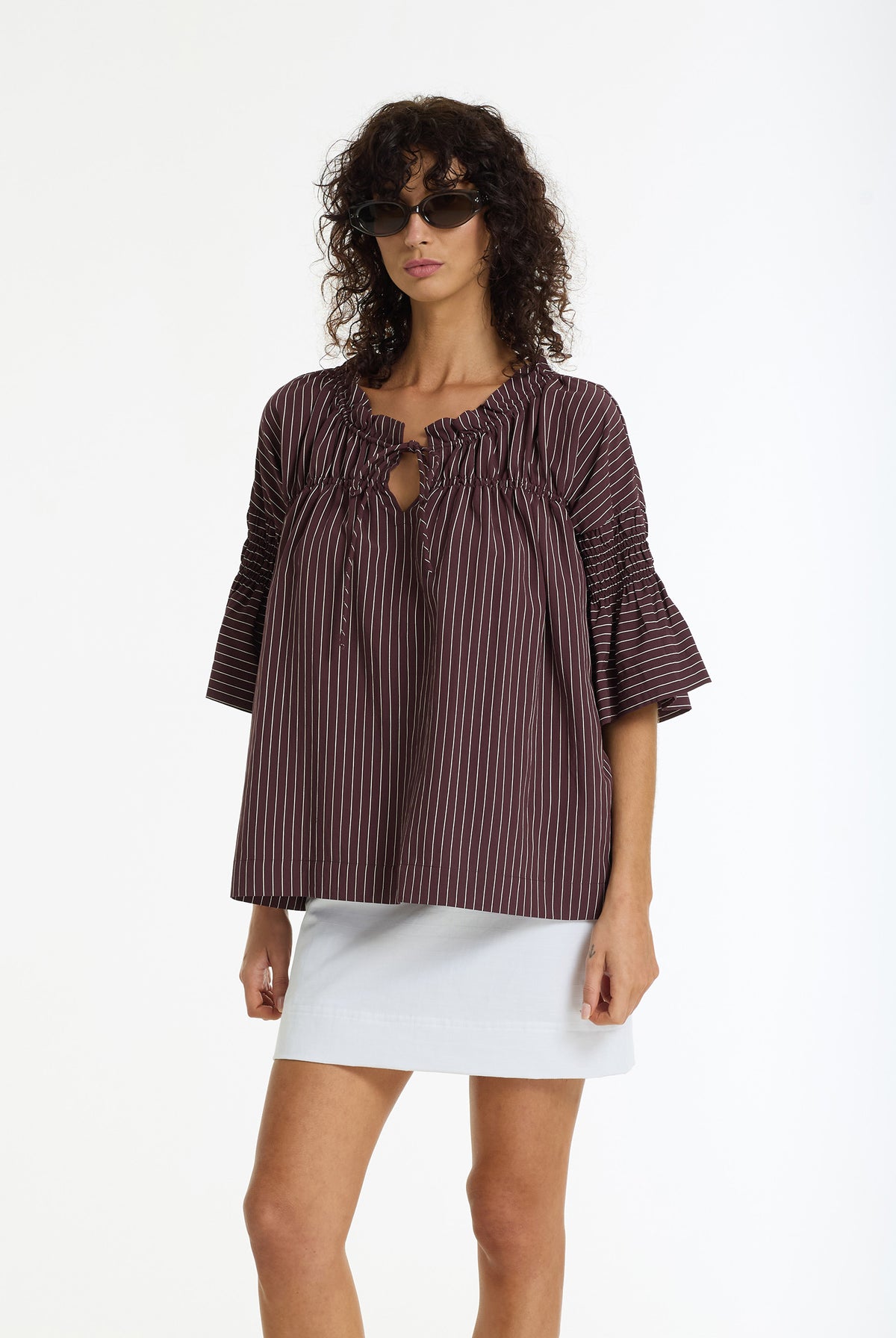 A model wearing a dark brown and black vertically striped blouse with a keyhole cutout and ruffled sleeves, paired with a white skirt, against a plain white background.
