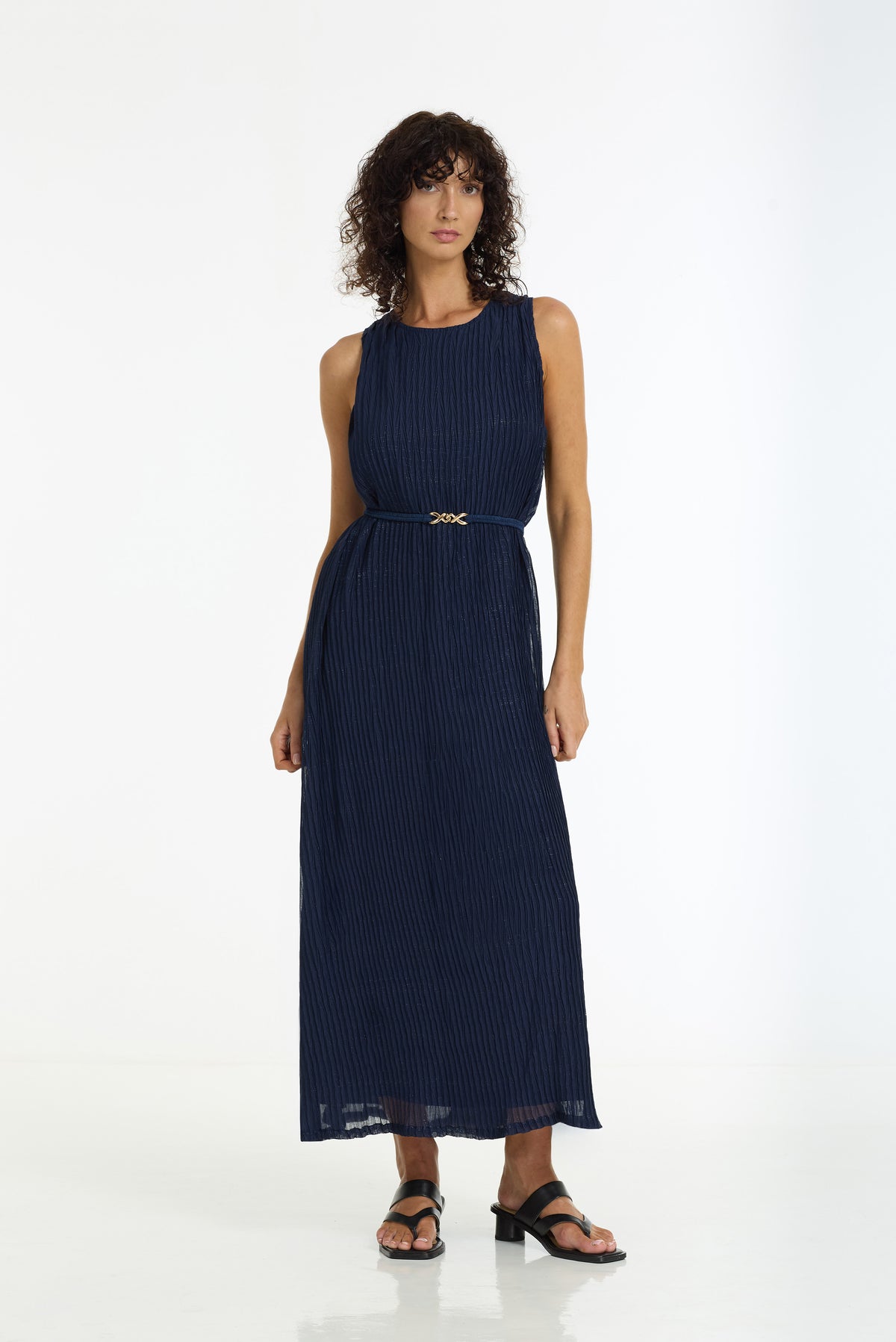 A woman modeling a sleeveless navy blue pleated maxi dress with a matching waist belt and a small gold cross detail, paired with black sandals, against a plain white studio background.