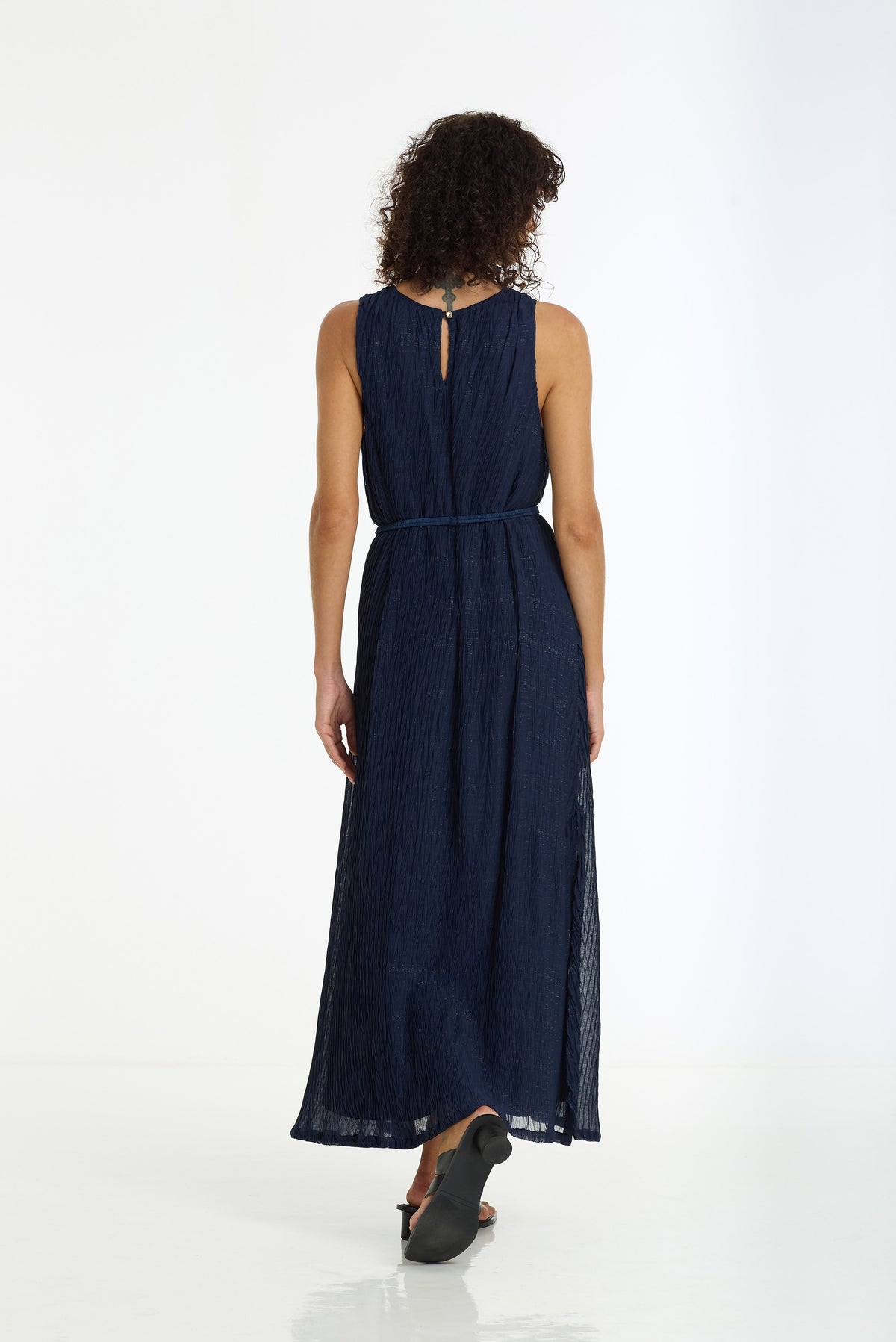 A woman viewed from behind wearing a sleeveless navy blue maxi dress with a tie-back neckline and a matching waist tie, standing against a clean white studio background.