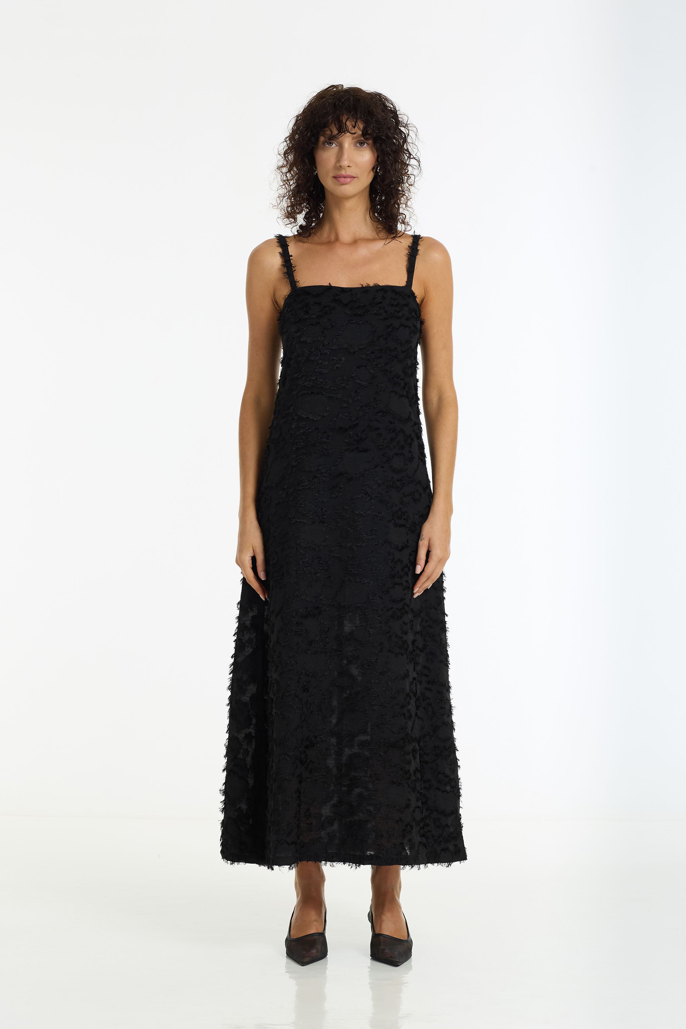 A woman wearing a Wildest Hearts Maxi Dress in a black textured fabric by BLAK, standing against a plain white studio background.