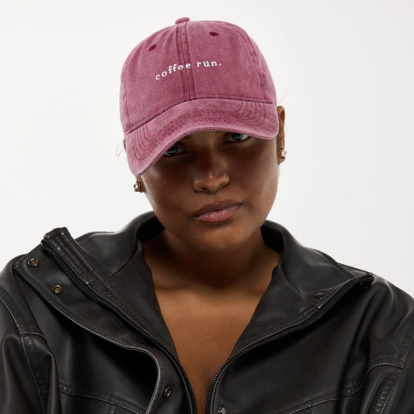A model wearing a dusty rose pink baseball cap with 'coffee run.' embroidered in white on the front, paired with a black leather jacket against a plain light gray background.