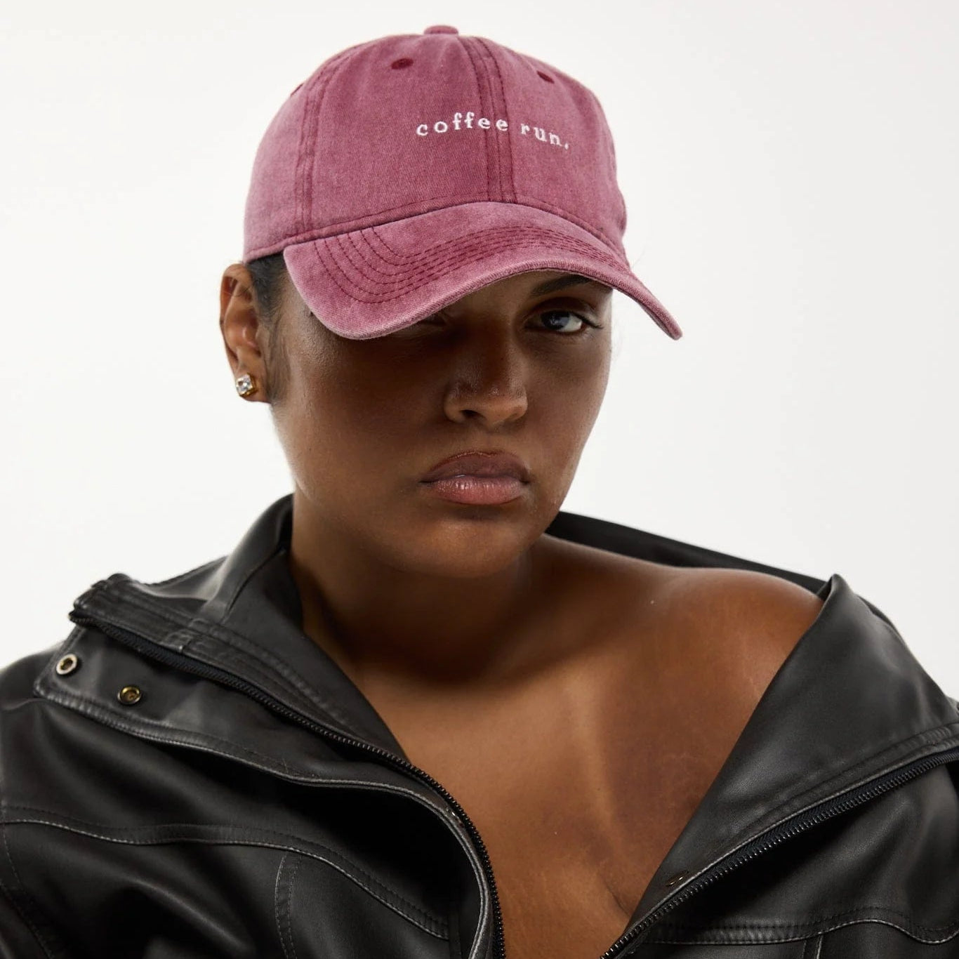 A close-up of a model wearing a dusty rose pink baseball cap with 'coffee run.' embroidered in white on the front, paired with a black leather jacket against a plain light gray background.