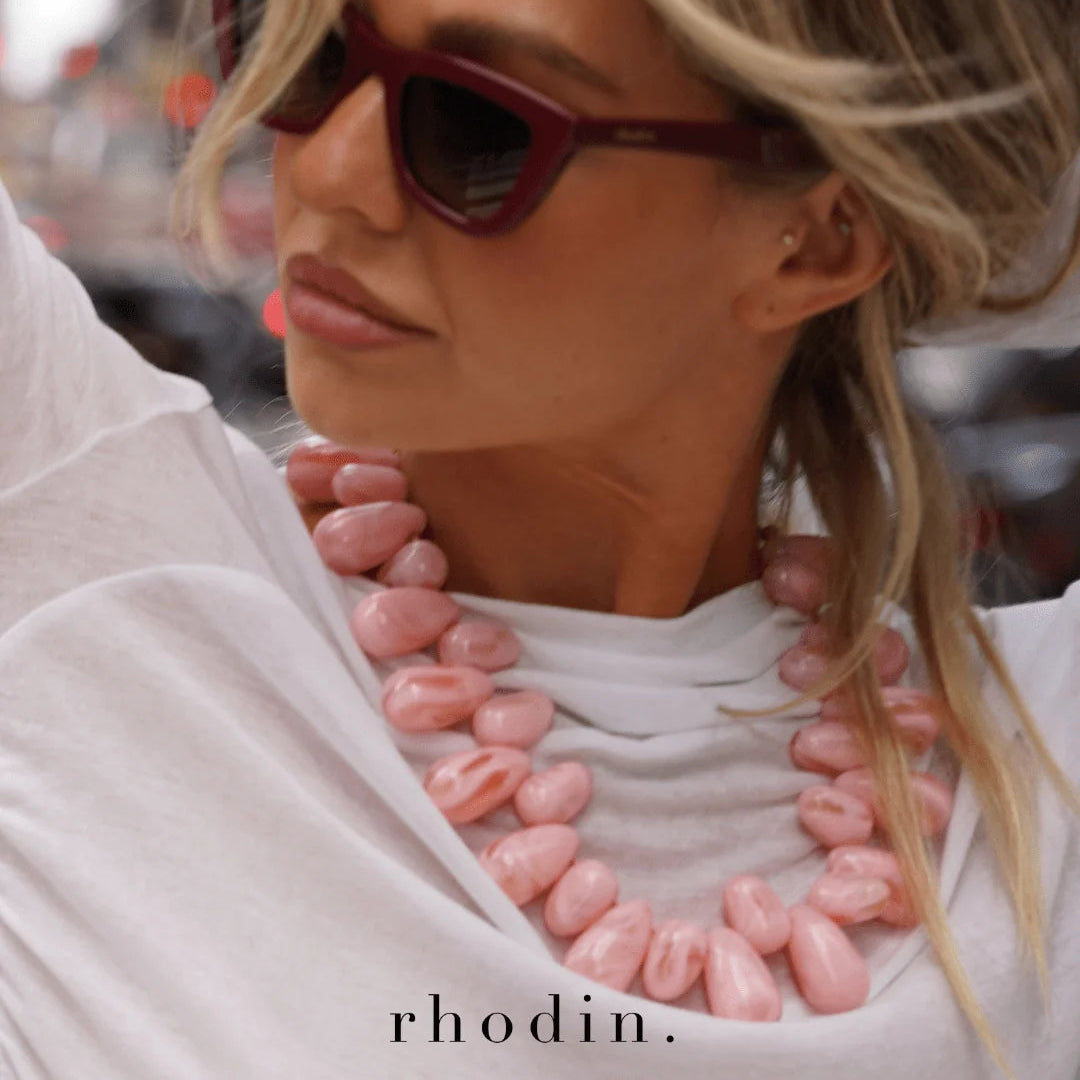 A close-up lifestyle shot of a woman wearing rhodin. chunky pink stone beads on a white t-shirt and dark red sunglasses.